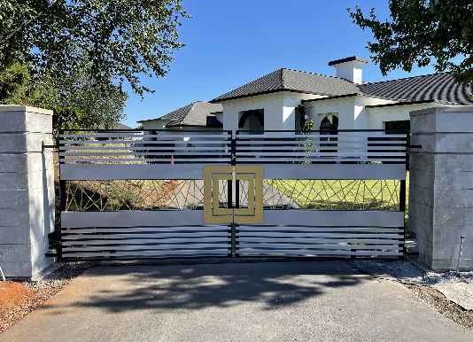 Metal gate with gold square handle in front of a modern house with a tiled roof and trees on side.