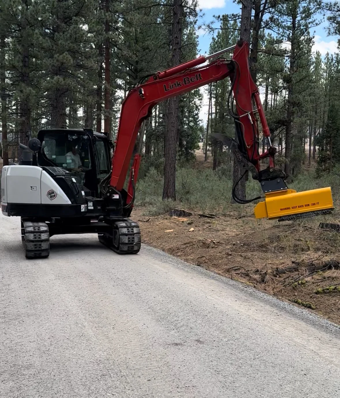 A small excavator digging into the dirt alongside a dirt road in a forested area.