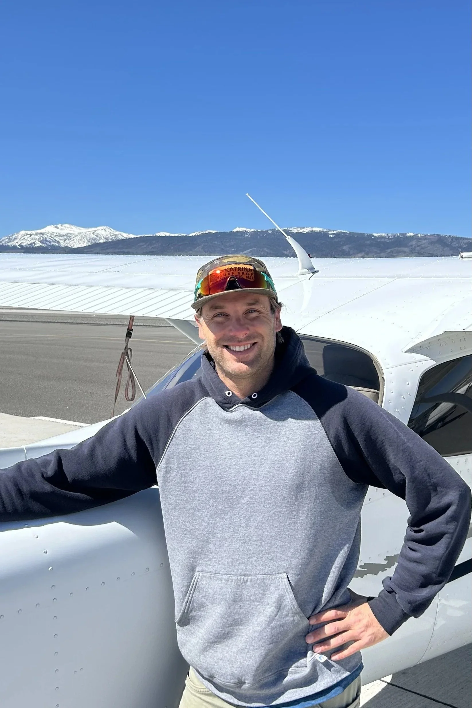 Man smiling in front of a small aircraft at an airport with snowy mountains and clear blue sky in the background.