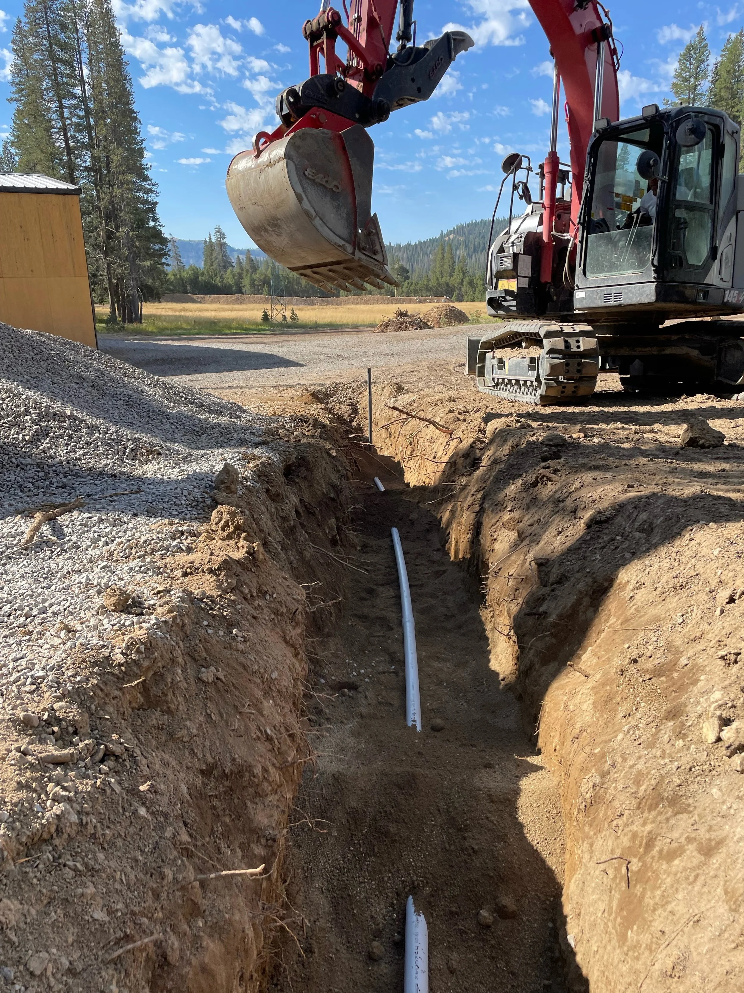 A construction site showing a trench with white PVC pipes installed in the ground, a red and black excavator with a large bucket, and a partly cloudy sky in the background.