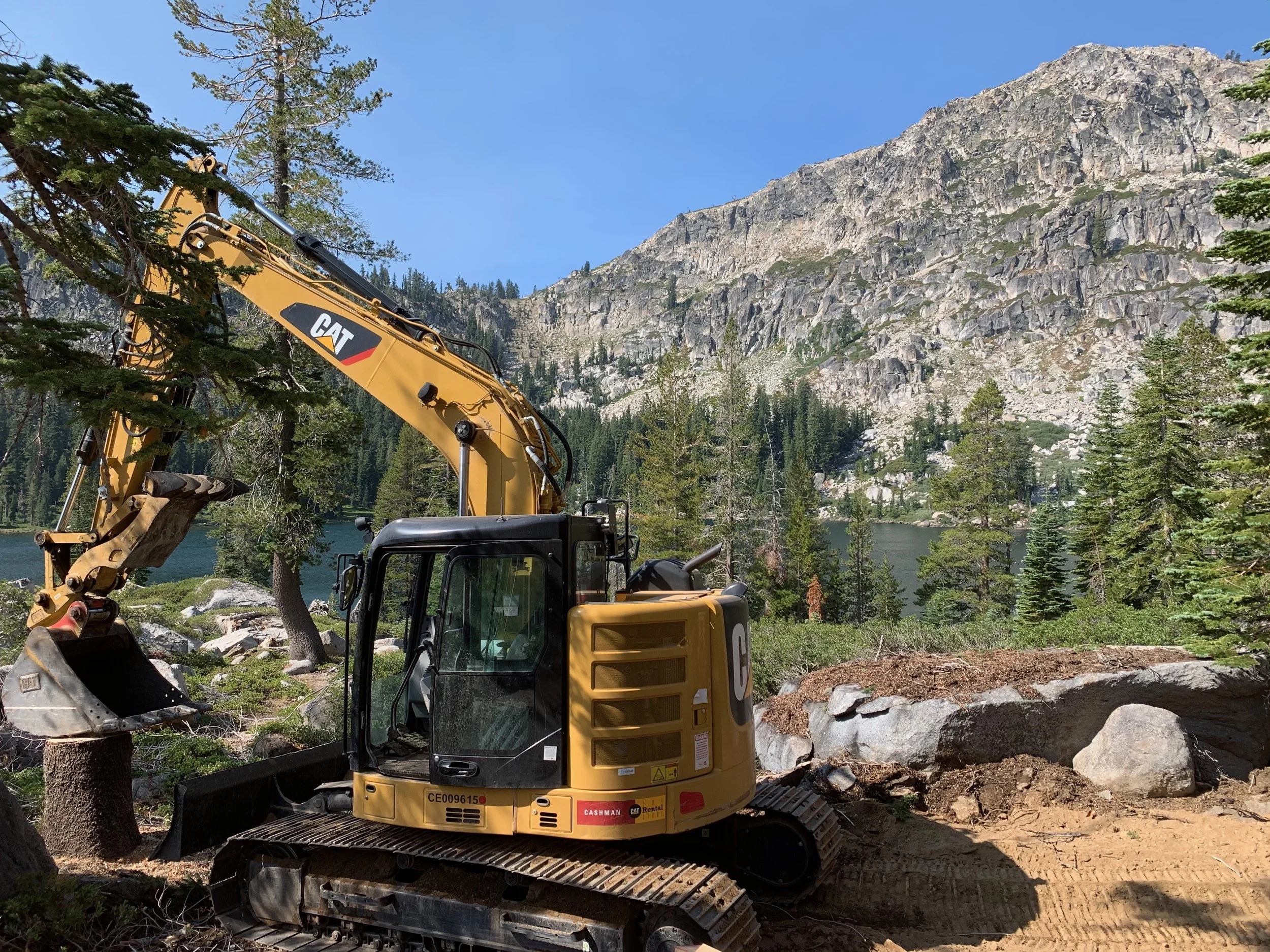 A yellow CAT excavator working on a dirt patch near a forested mountain lake, with trees, rocks, and a mountain in the background.