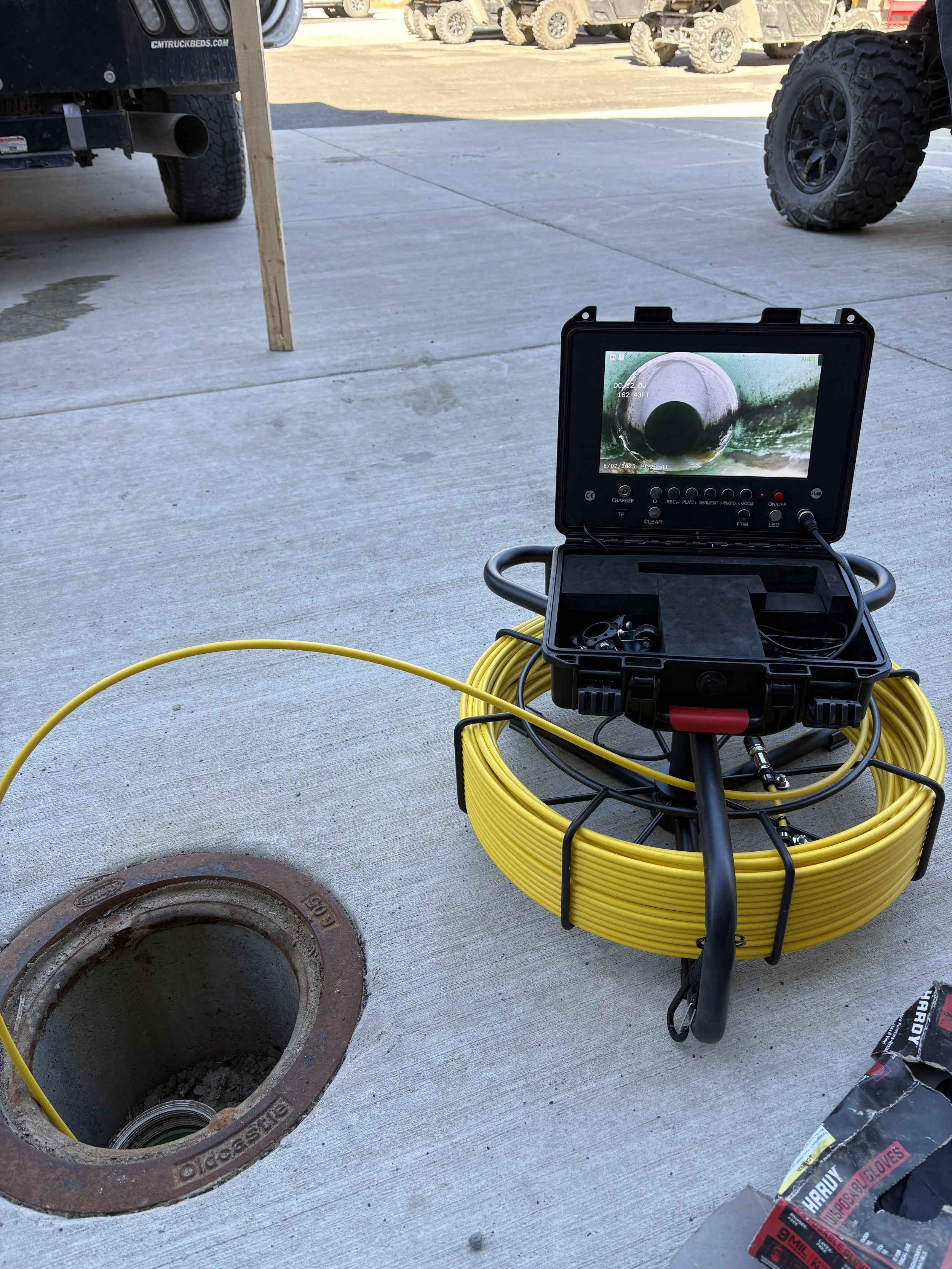 A pipe inspection camera setup with a screen displaying the interior of a pipe, positioned next to an open sewer drain in a concrete surface.