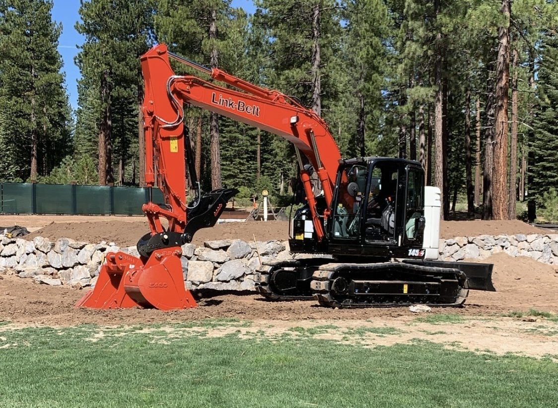 A red and black compact excavator with the brand Link-Belt is working on a construction site with dirt and rocks, surrounded by tall pine trees.