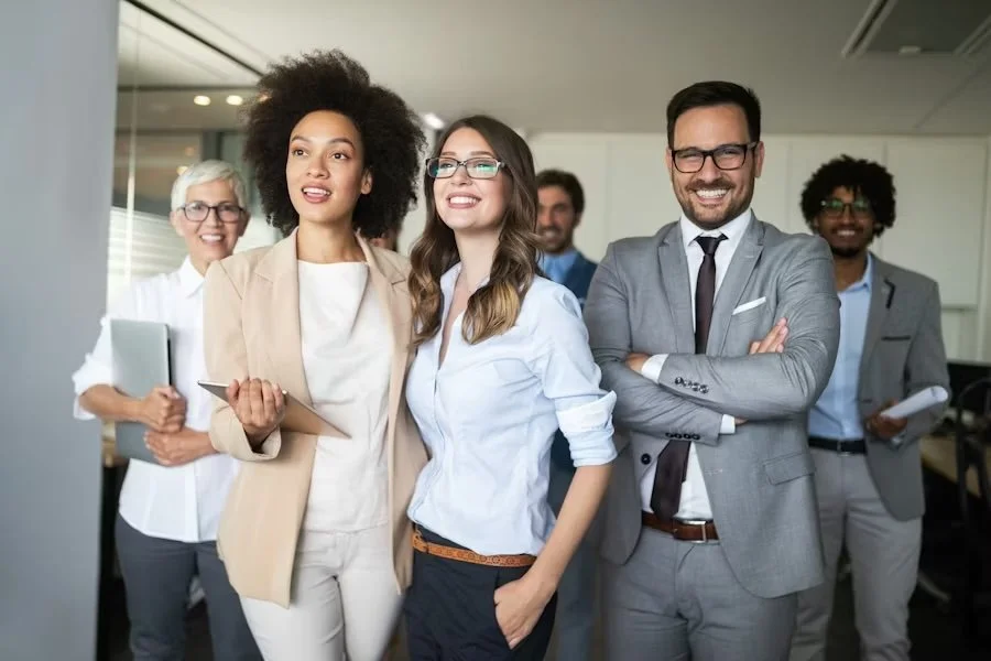 Group of six diverse professionals in an office, smiling and looking towards a presentation or screen, dressed in business attire.