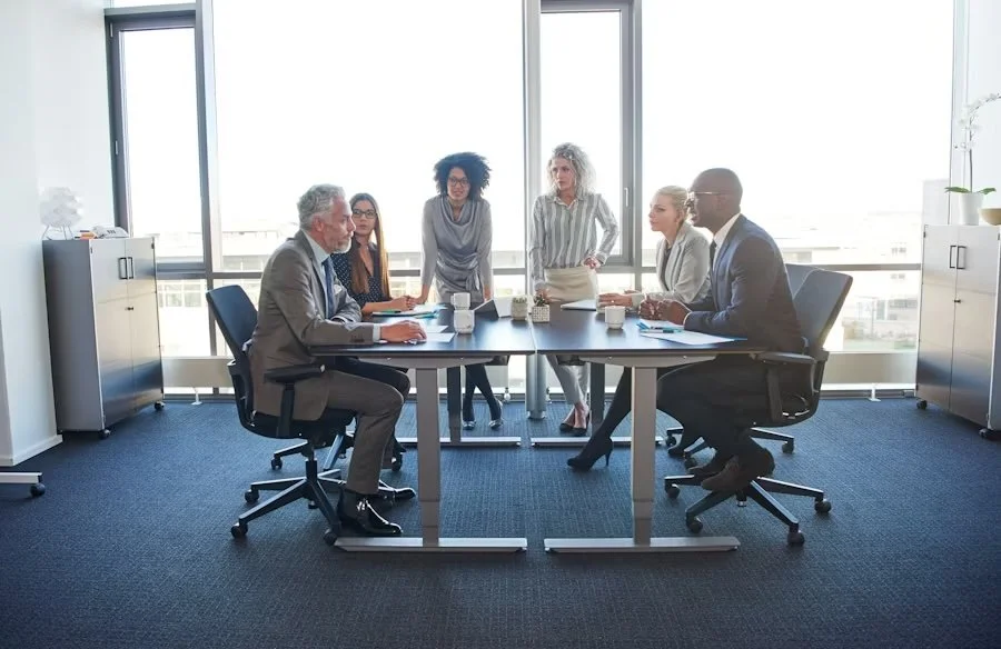 Business meeting with six diverse professionals around a conference table in a modern office with large windows.