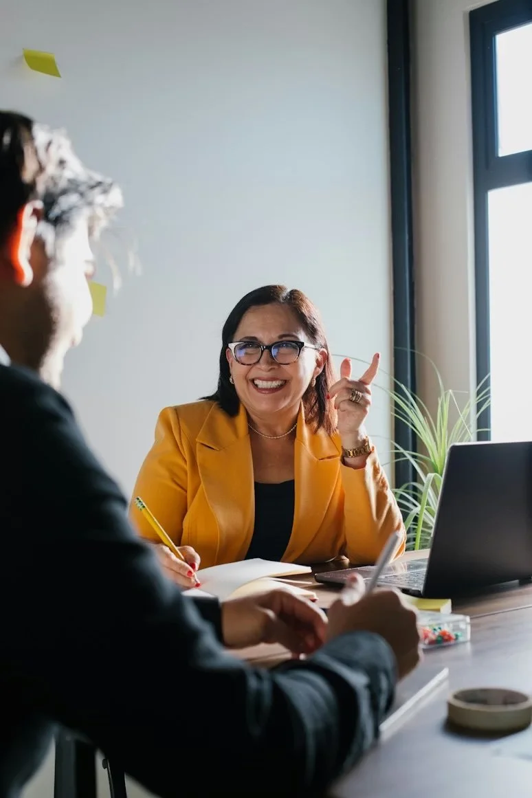 Woman in yellow blazer and glasses smiling and talking during a business meeting in an office.