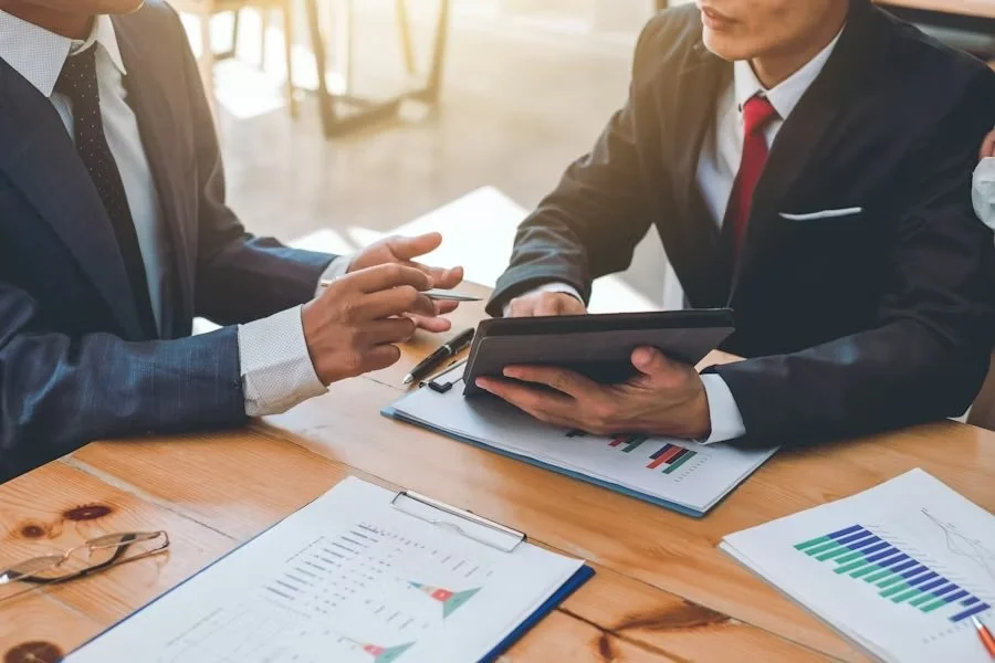 Two businessmen in suits having a discussion at a wooden desk with financial charts and documents.
