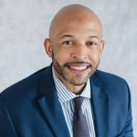 A professional man with a bald head and a beard, dressed in a blue suit, white shirt, and blue tie, smiling against a light background.