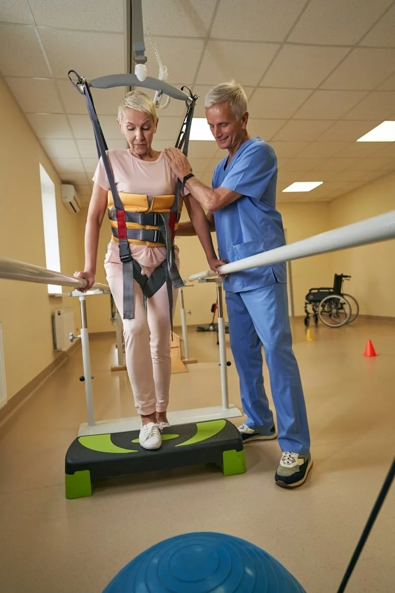 A senior woman with short gray hair using a harness and parallel bars for support during a rehabilitation session, assisted by a healthcare professional in blue scrubs, in a therapy room with wheelchair and exercise equipment visible.