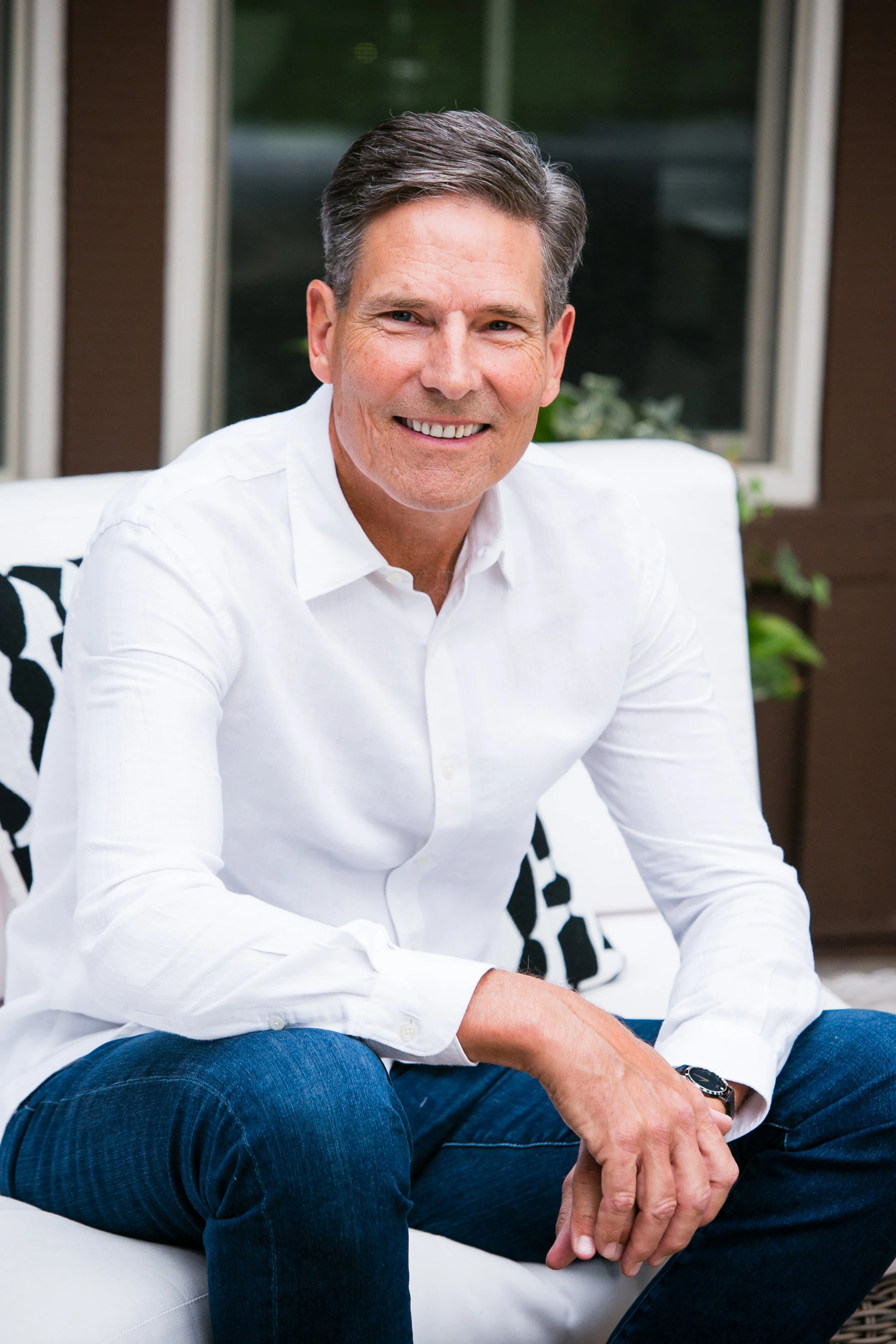 A middle-aged man with dark hair, wearing a white button-up shirt and jeans, sitting on a white outdoor sofa. Behind him is a window with green foliage visible outside.