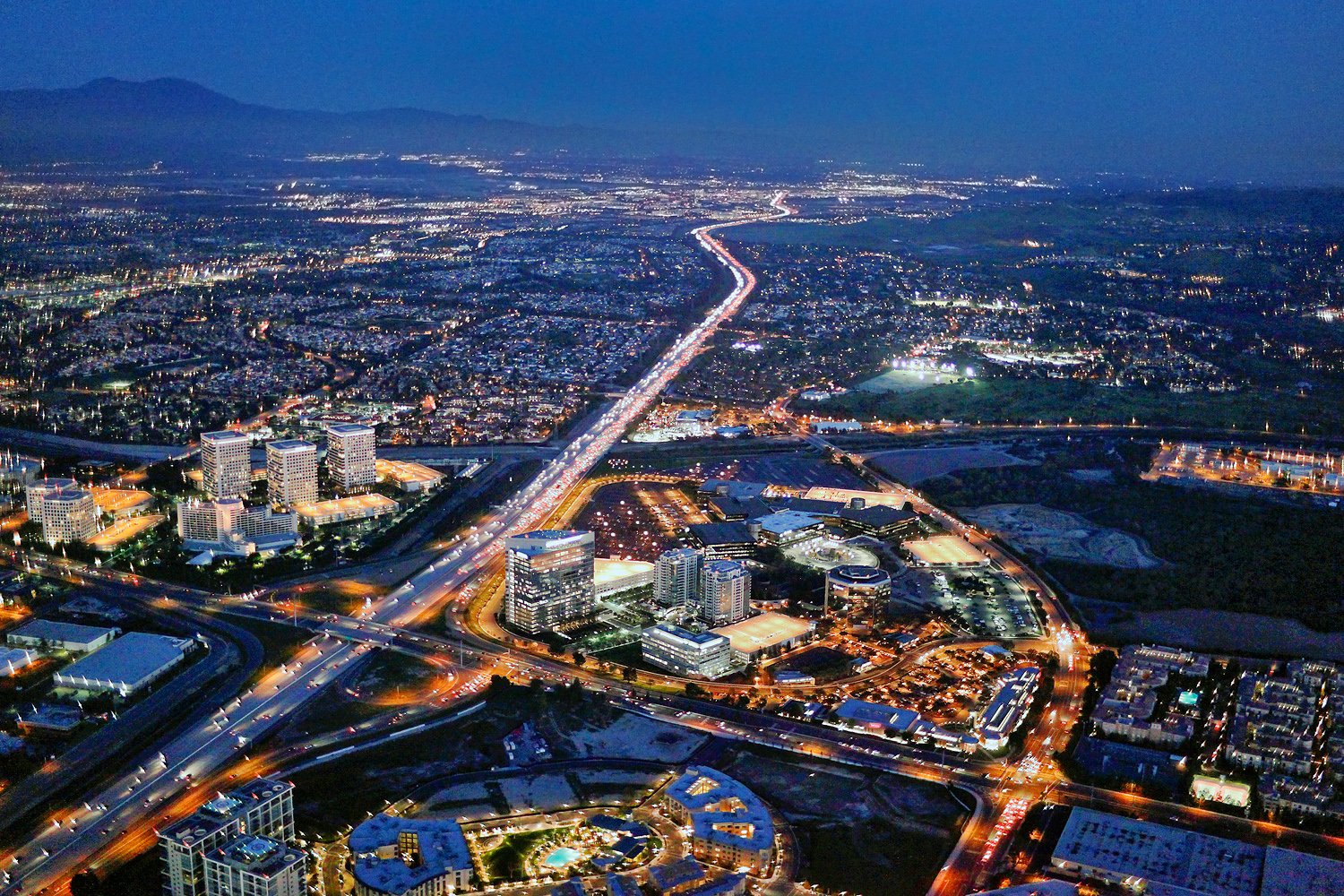 Aerial view of a city at night, showing illuminated roads, buildings, and highways with mountains in the background.