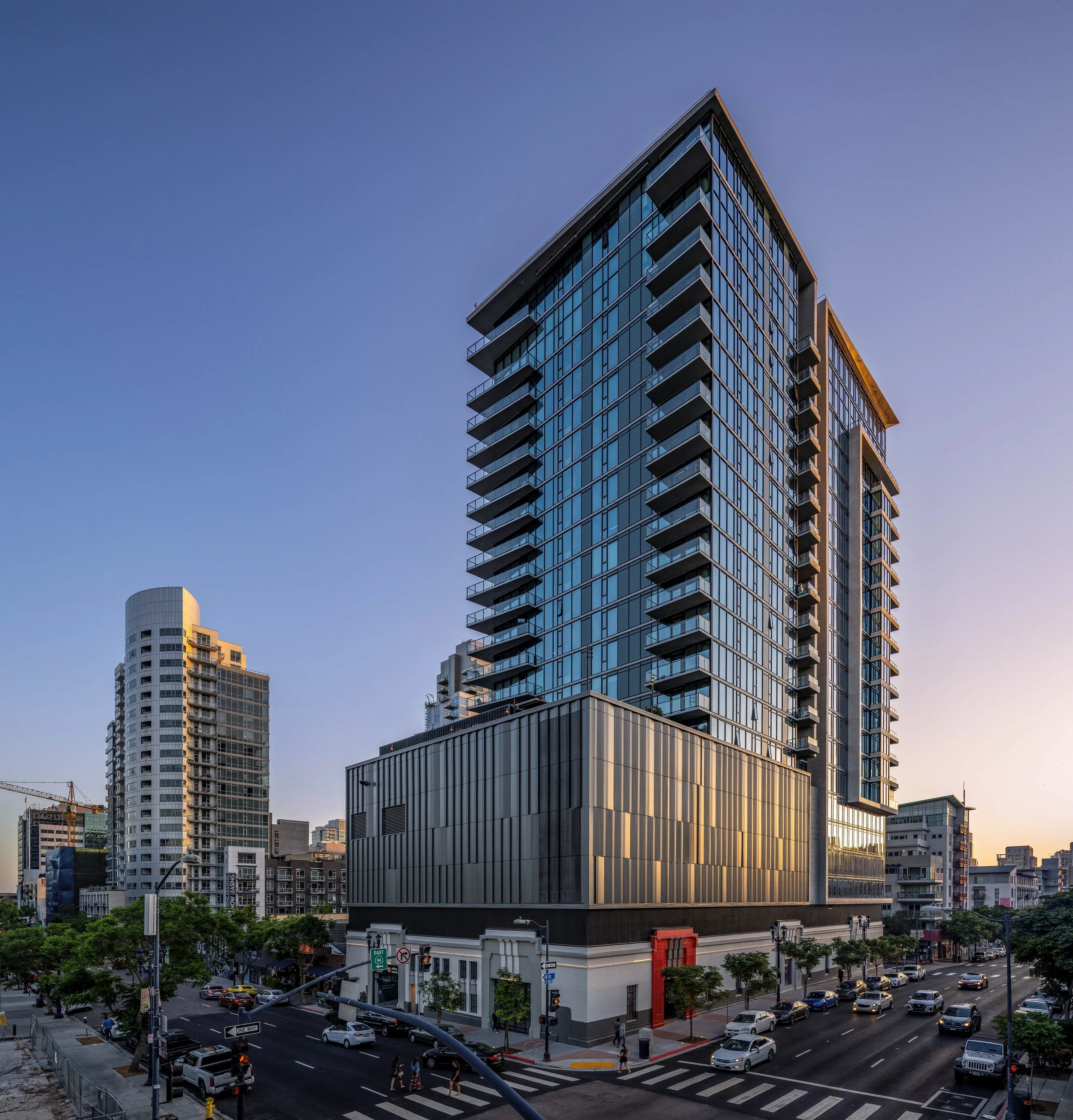 A modern high-rise office or apartment building with large glass windows and multiple balconies, situated on a city street with cars and pedestrians, under a clear blue sky during dusk or dawn.
