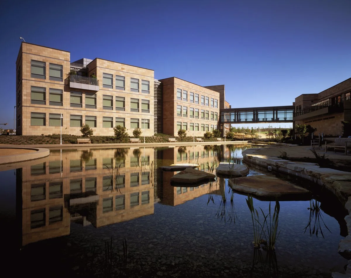 Modern office building with multiple windows, a glass skywalk connection, and a reflecting pond in the foreground, under a clear blue sky.