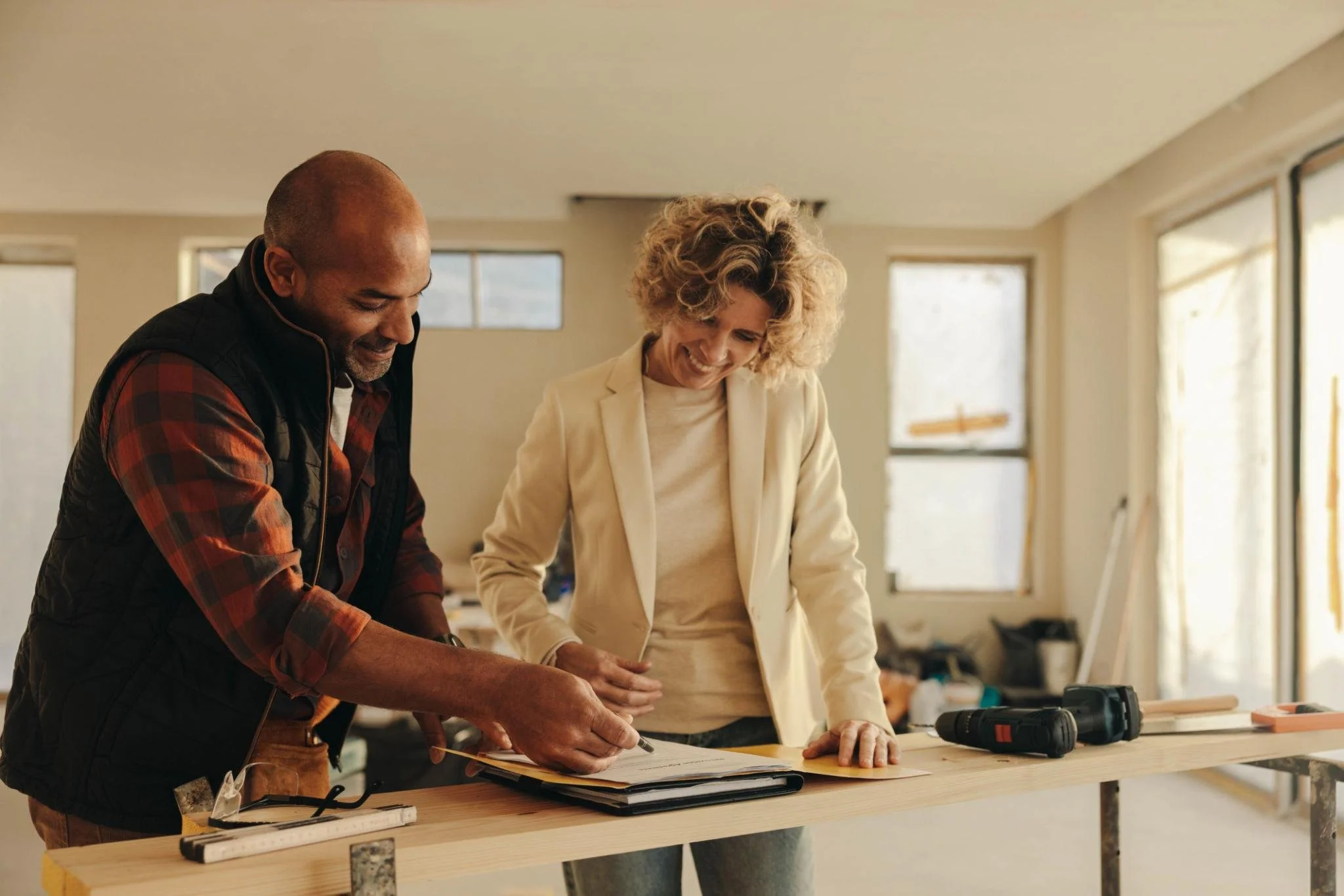 Two smiling people, a man and a woman, look at documents on a workbench inside a bright, unfinished room. The man is signing a paper while the woman watches.