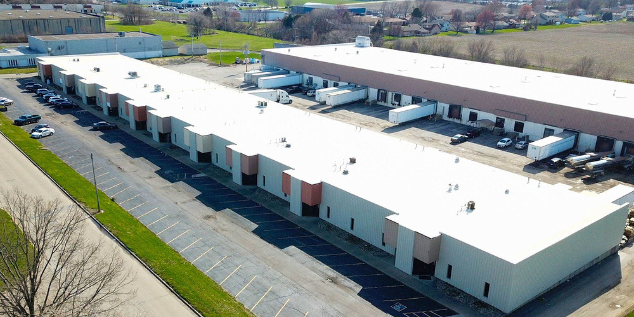 Aerial view of a large commercial building with a white roof, parking lot, trucks, and surrounding greenery.