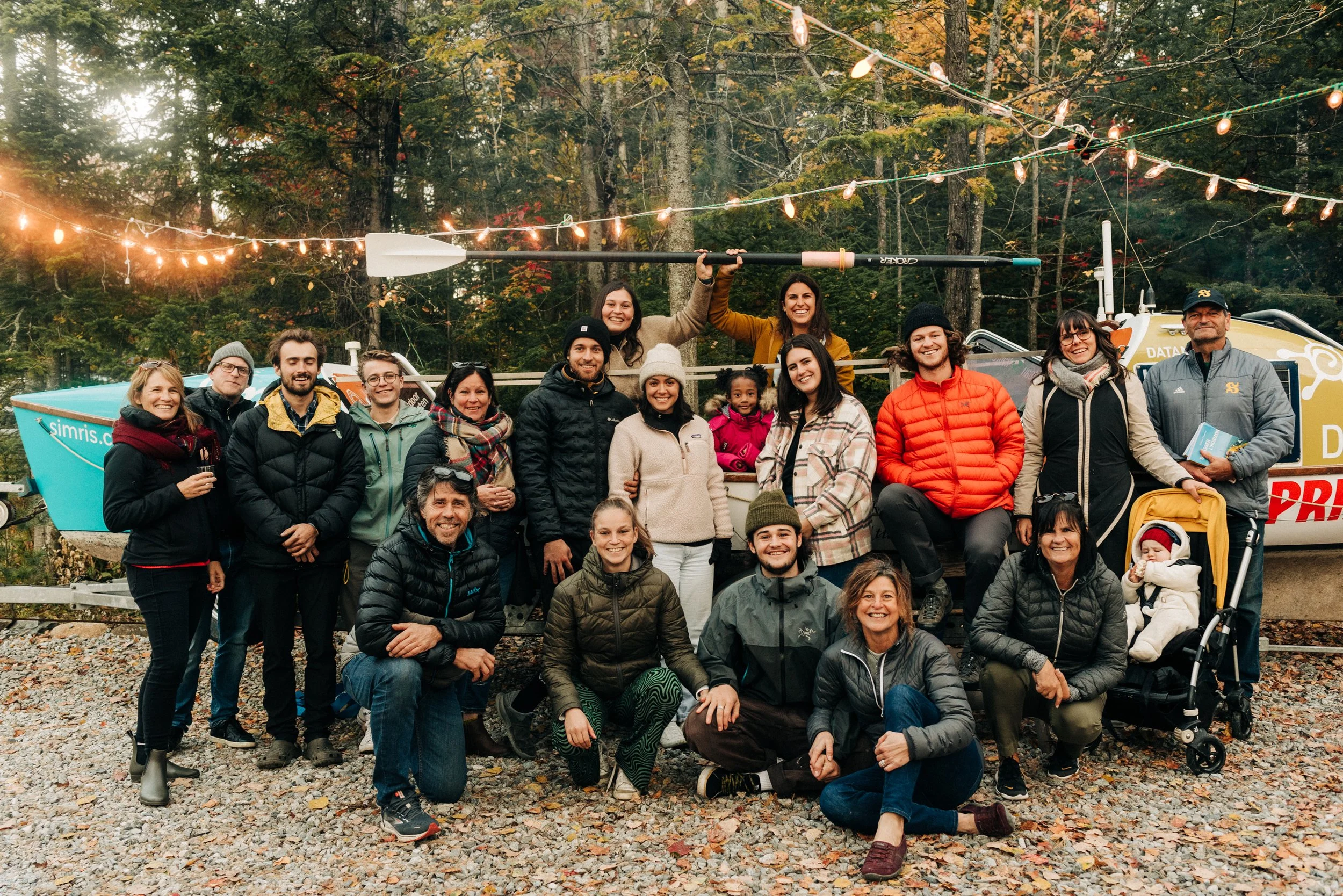 Groupe d'une vingtaine de personnes posant pour une photo en forêt avec un bateau en arrière-plan, décorée de guirlandes lumineuses