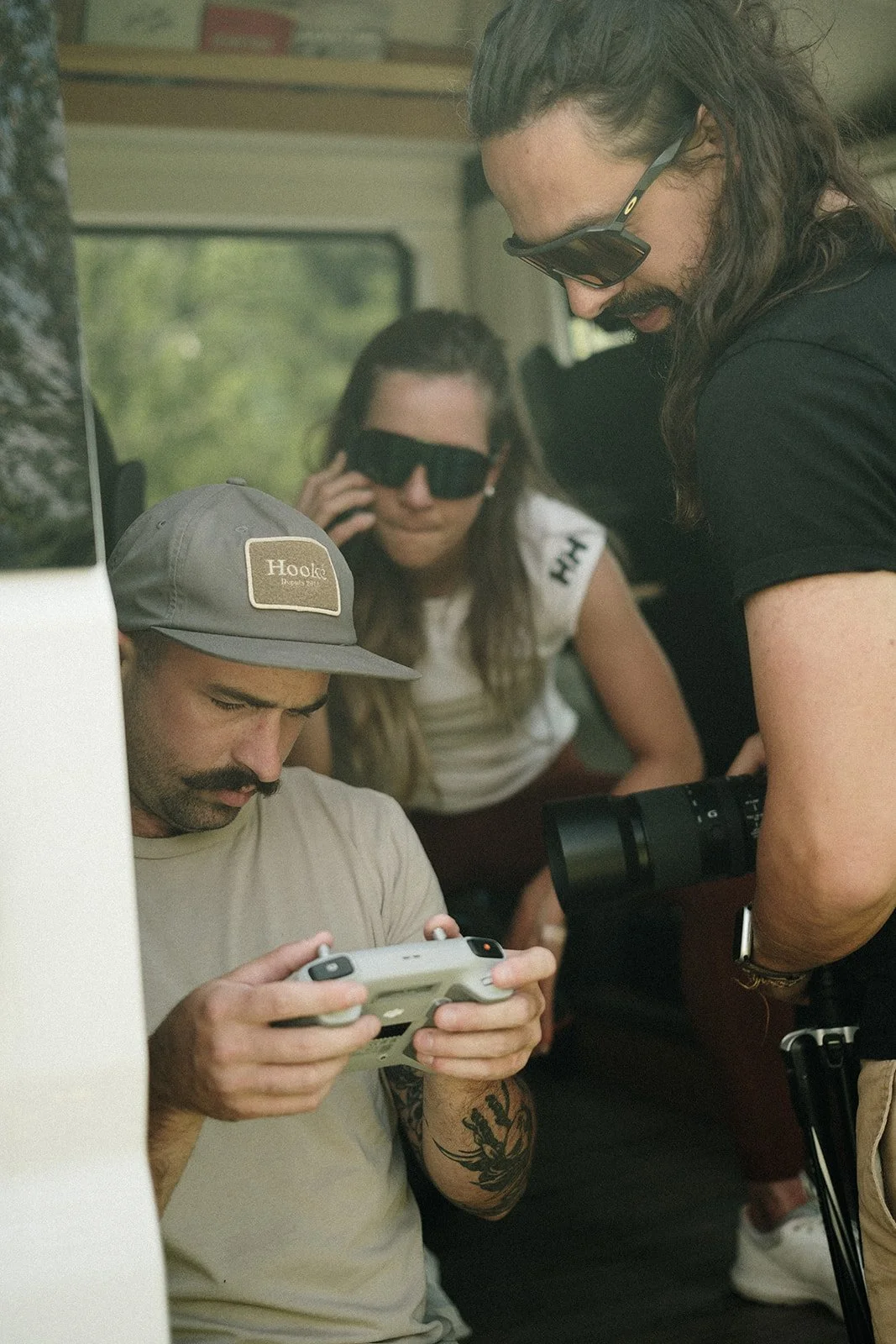 Trois personnes regardant un appareil électronique, deux portant des lunettes de soleil et une portant une casquette, à l'intérieur d'un véhicule.