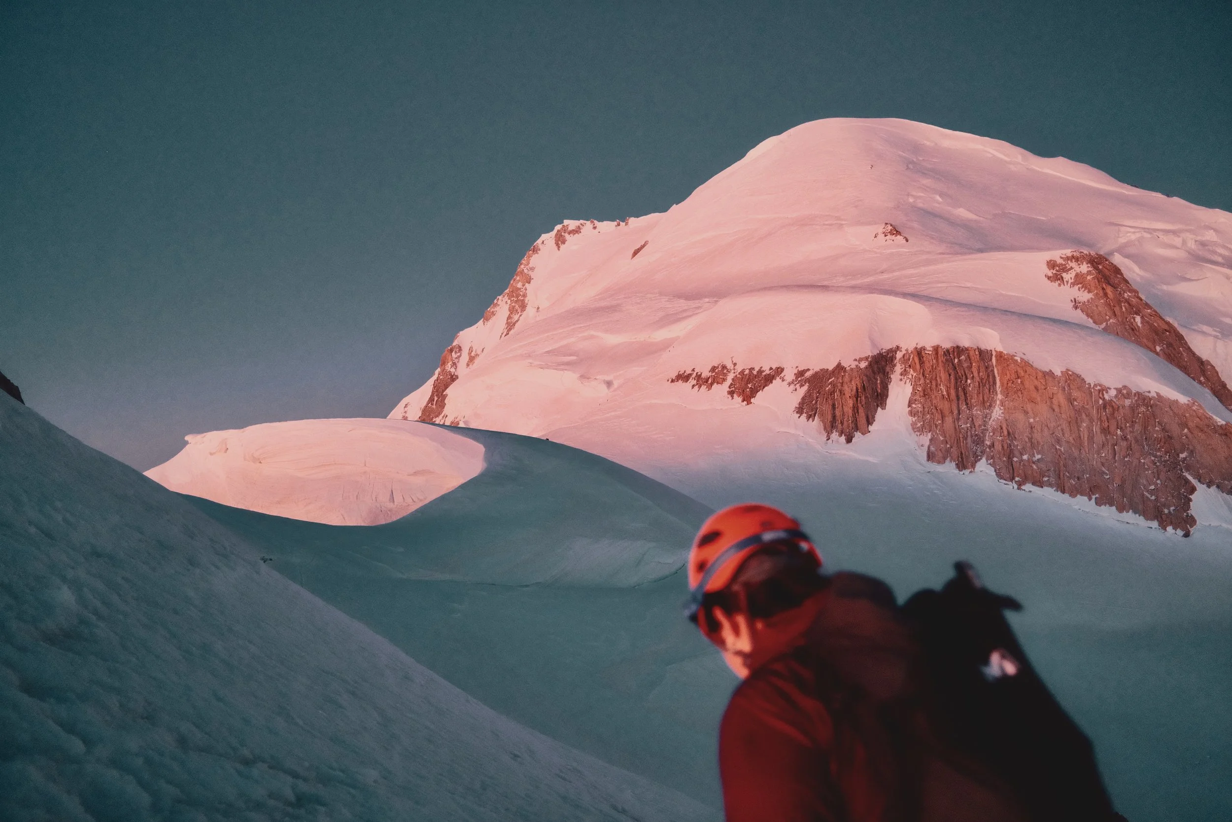 Un alpiniste portant un casque orange, regardant vers un sommet de montagne enneigé au coucher du soleil.