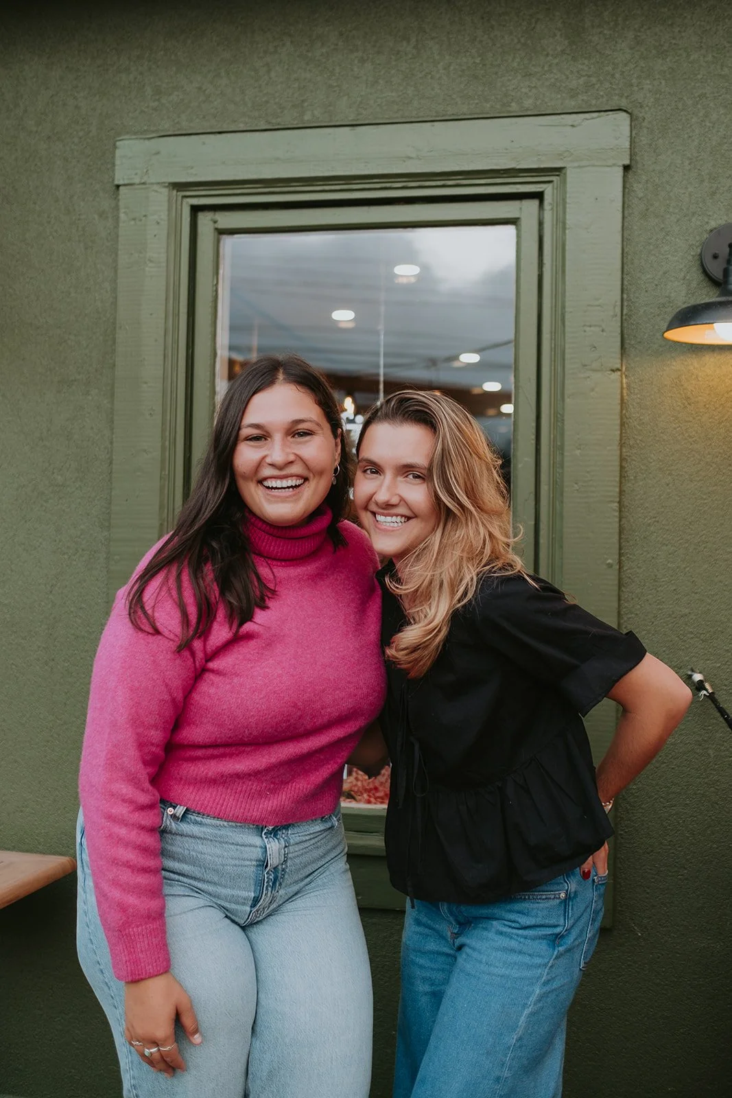 Deux femmes souriantes debout devant une fenêtre verte, portant des jeans, l'une en pull rose et l'autre en chemise noire, en intérieur