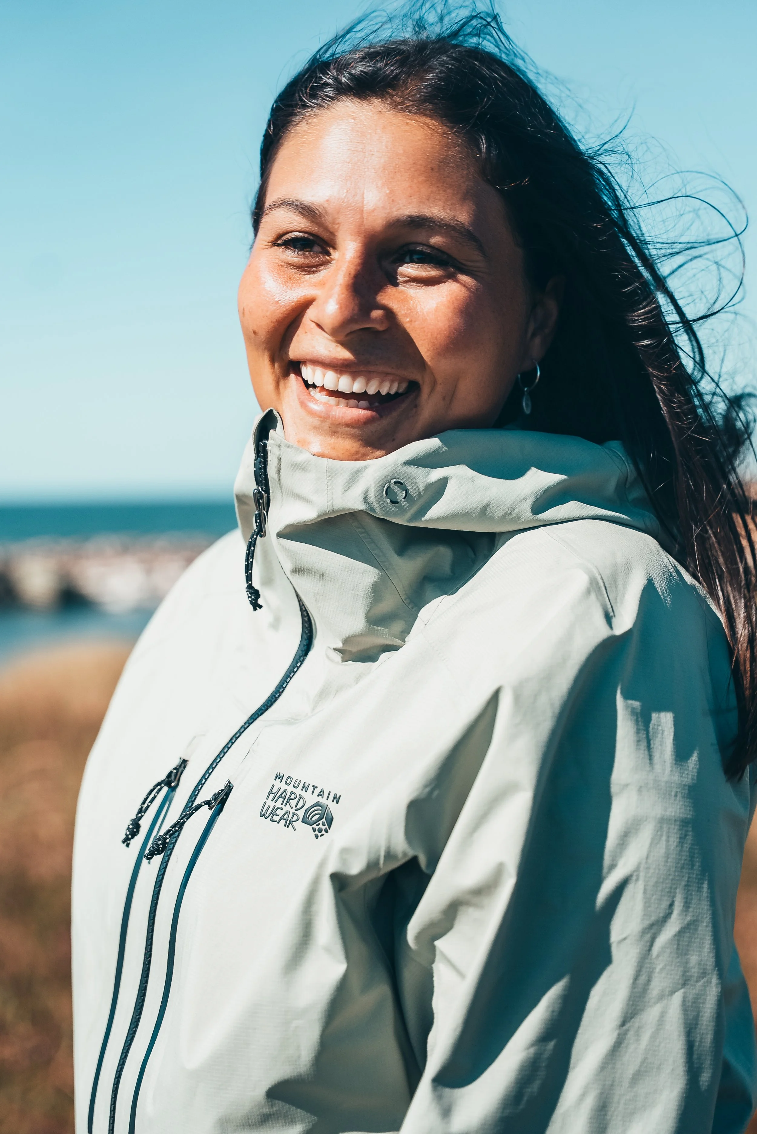 Anabelle Guay portant un manteau 'Mountain Hard Wear', à l'extérieur avec un ciel bleu et la mer en arrière-plan.