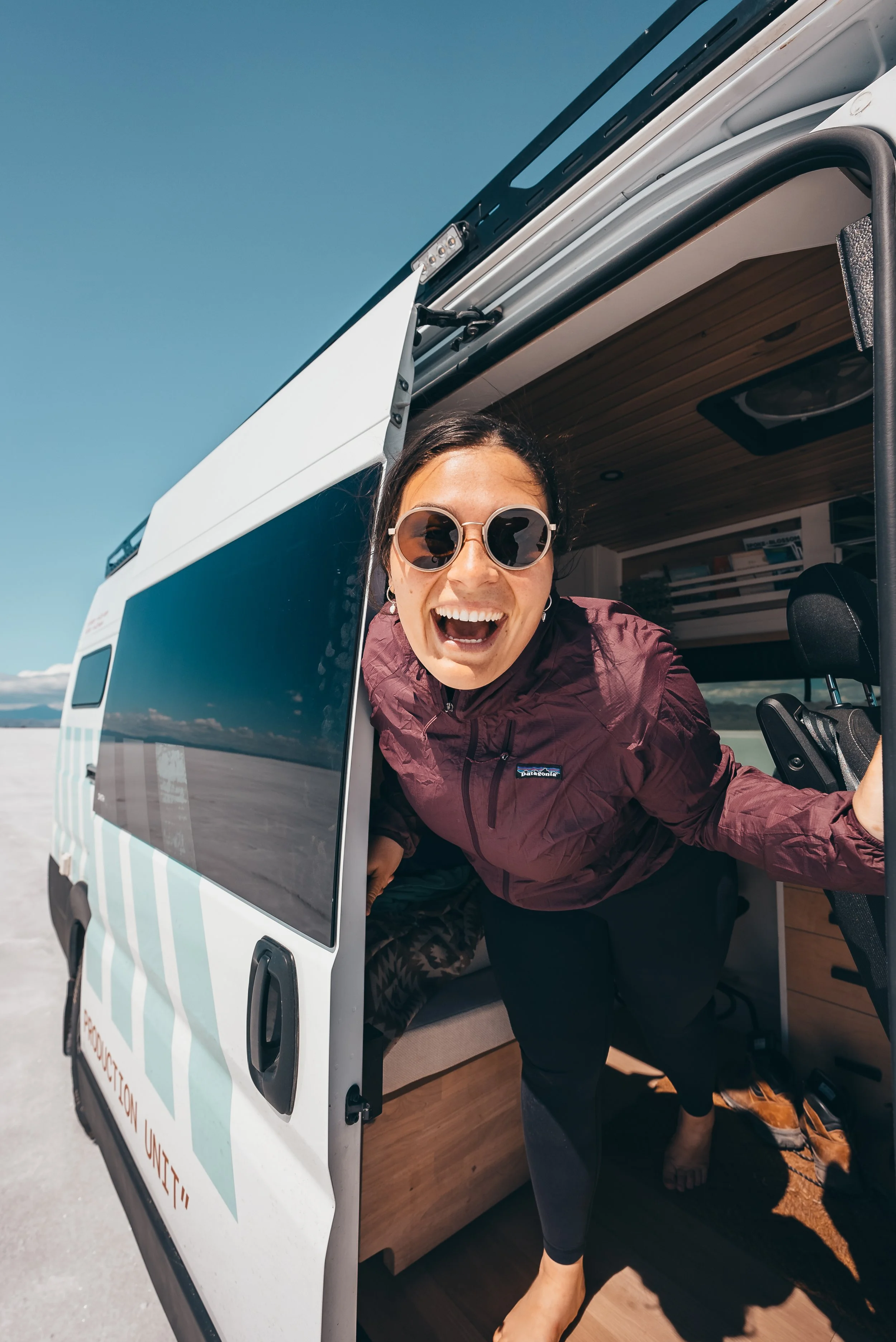 Femme souriante portant des lunettes de soleil, sortant d’un van blanc avec intérieur en bois, dans un paysage à ciel bleu.