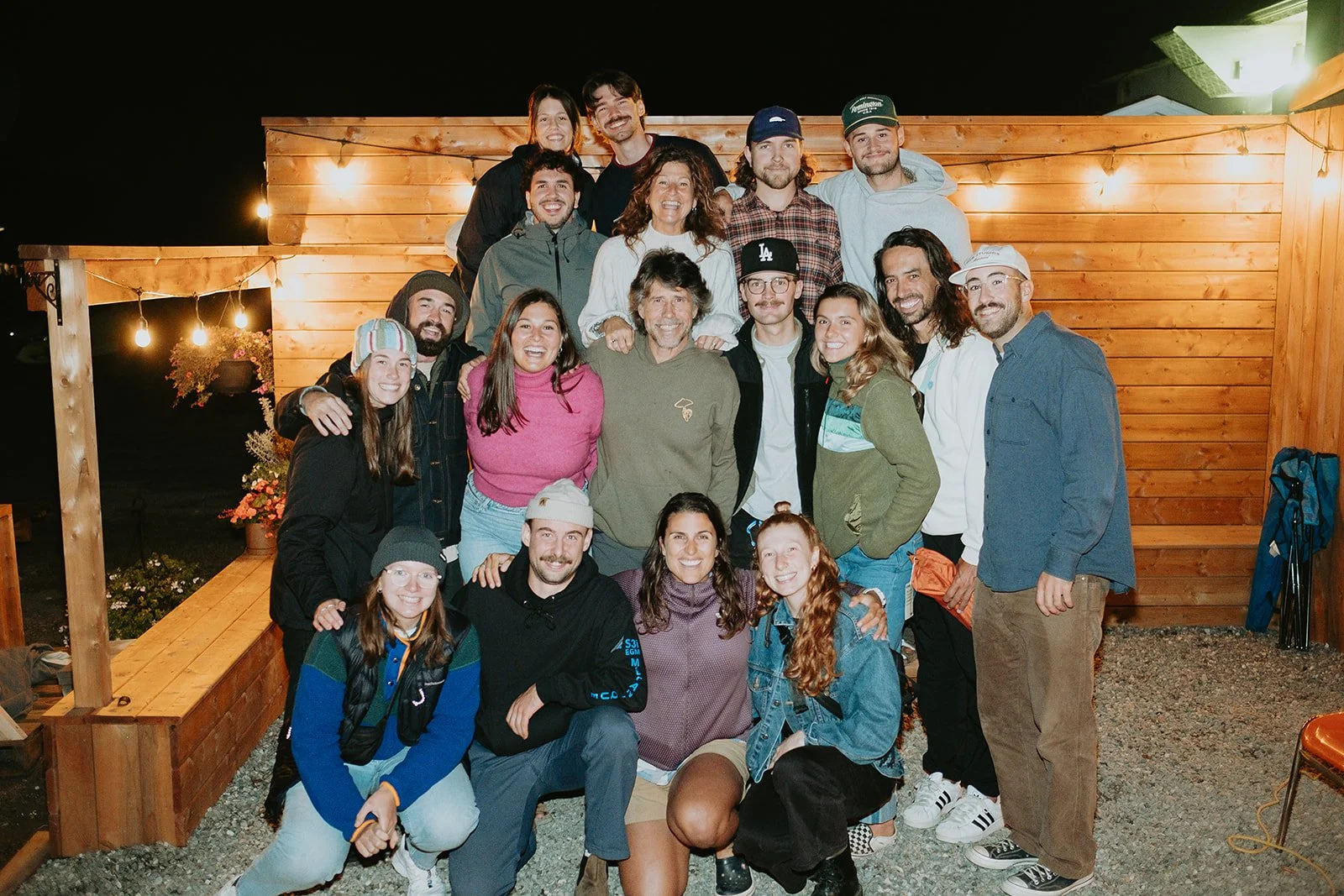 groupe de personnes posant ensemble devant une structure en bois, lors d'une soirée, ambiance chaleureuse avec éclairage de guirlandes