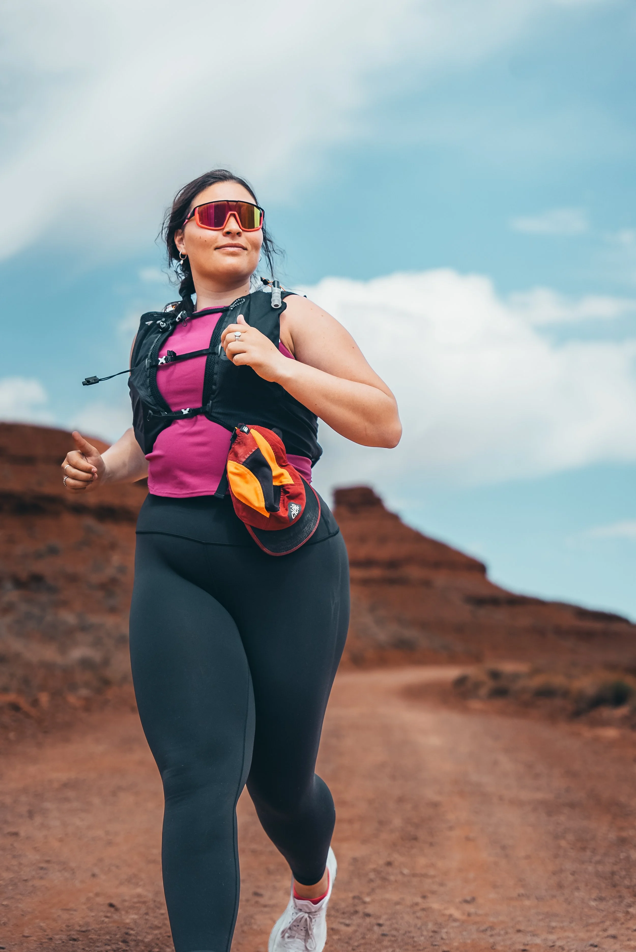 Femme courir dans un environnement désertique, portant un débardeur rose, une veste sans manches, des leggings noirs, des lunettes de soleil et un sac à dos