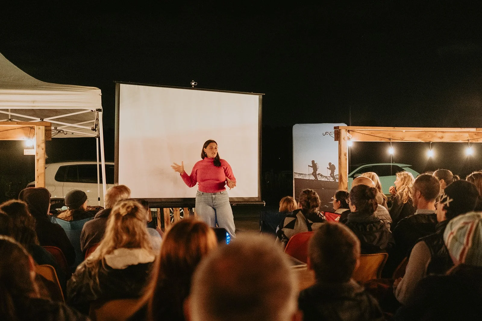 Une femme parle devant un public lors d'une projection en plein air la nuit, avec un écran blanc derrière elle et des décorations en bois avec des lumières