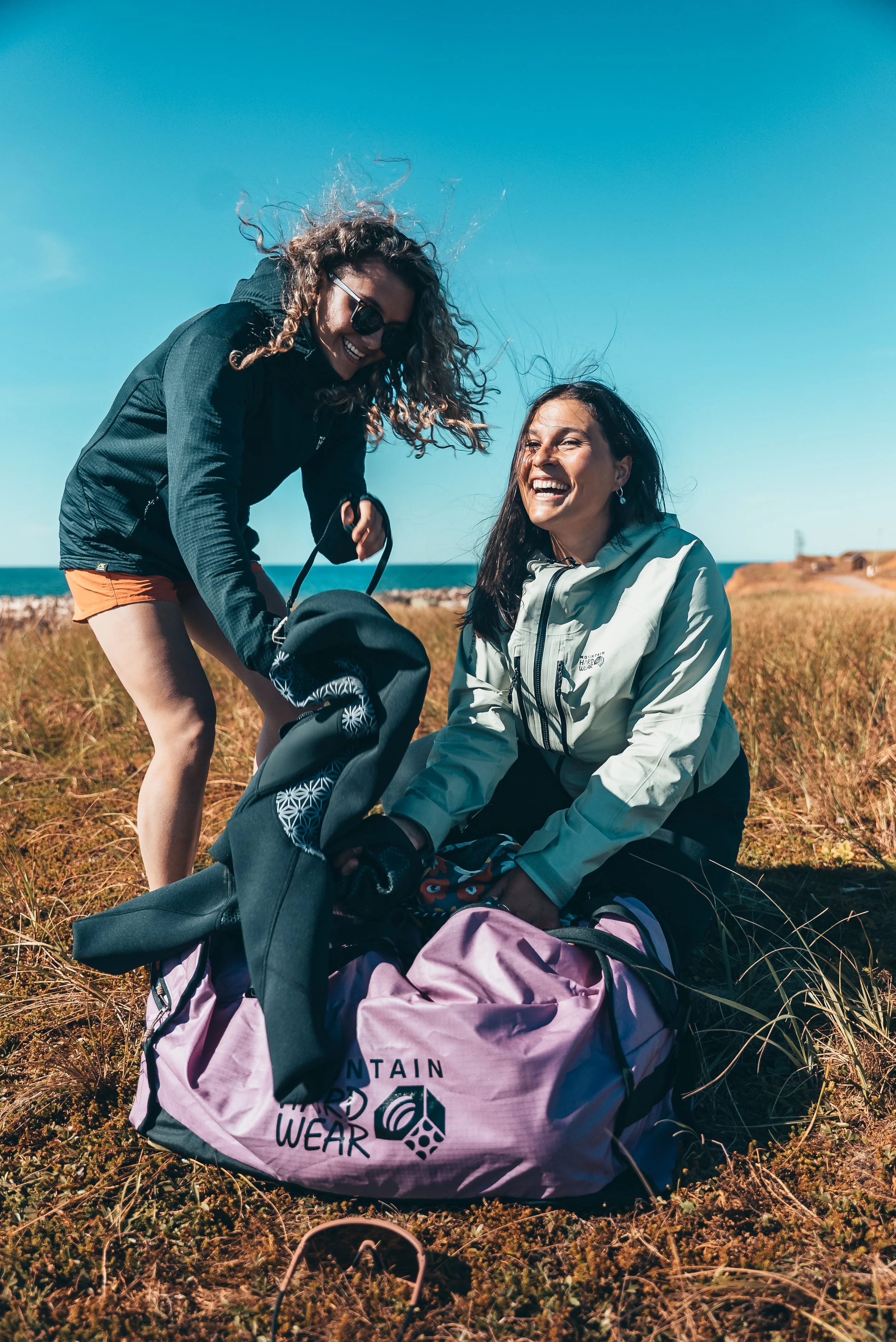 Deux femmes souriantes dans un paysage côtier, préparant leurs sacs à dos pour une randonnée ou une excursion en plein air, avec un ciel bleu clair.