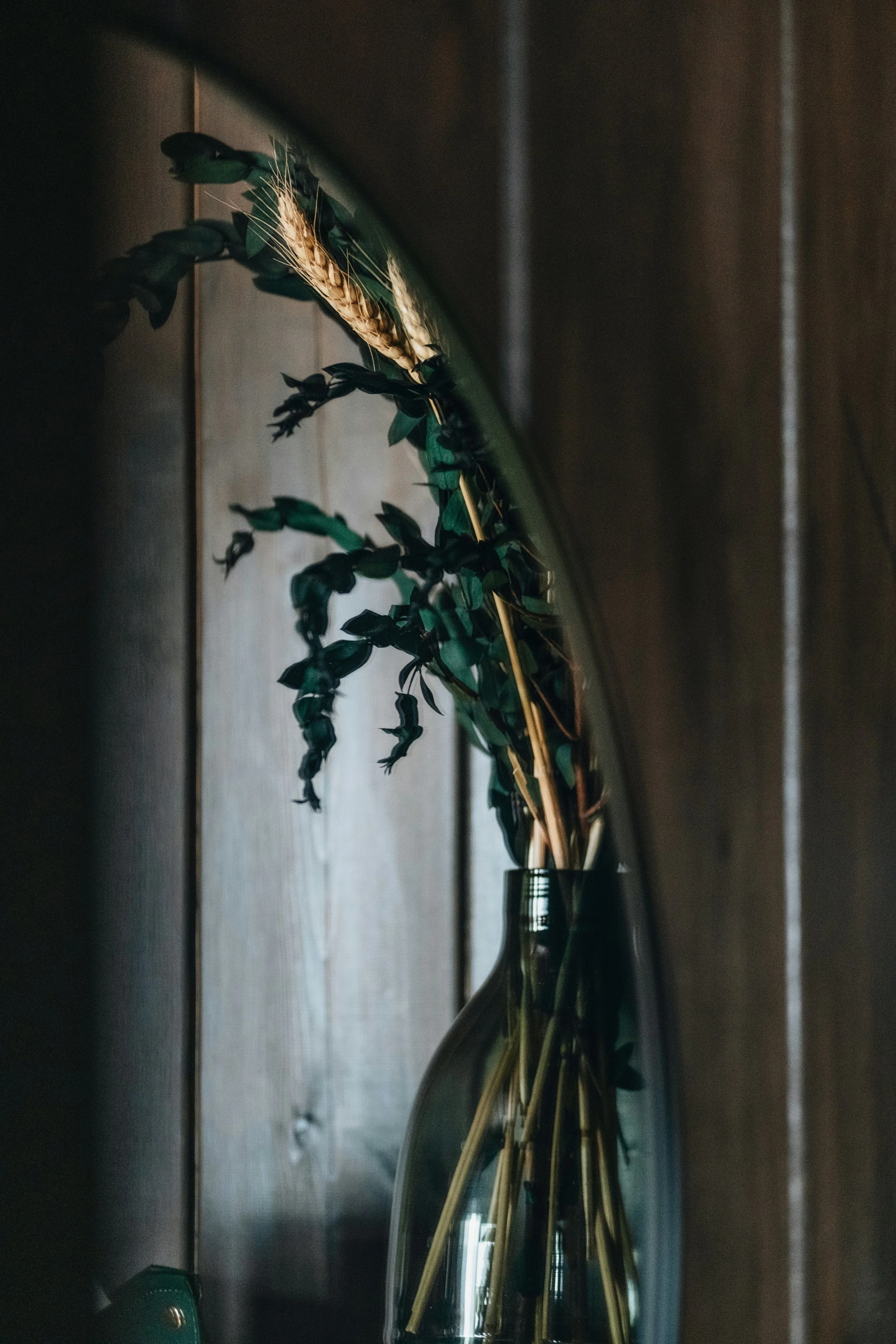 Close-up of a decorative mirror reflecting dried wheat stalks in a brass vase, placed on a wooden surface.
