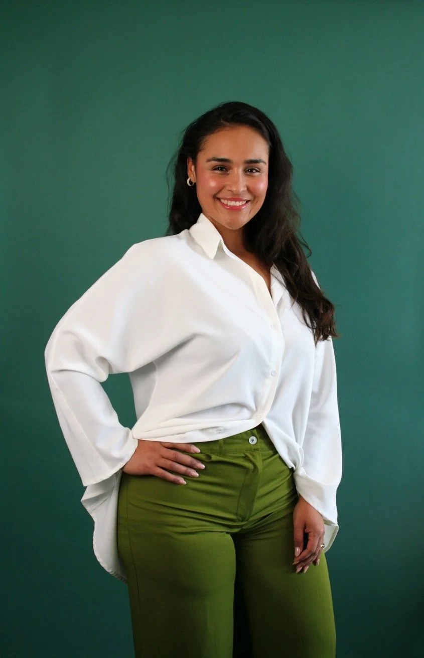 A woman with dark, wavy hair and medium skin tone smiles at the camera while standing against a solid teal background. She is wearing a white, long-sleeved blouse and olive green pants.