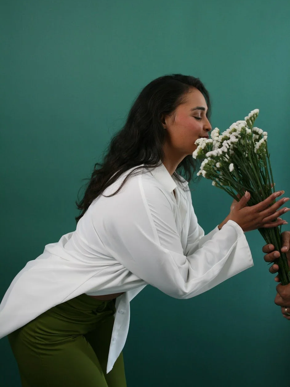 A woman with dark hair and wearing a white shirt is smelling a bouquet of white flowers against a green background.