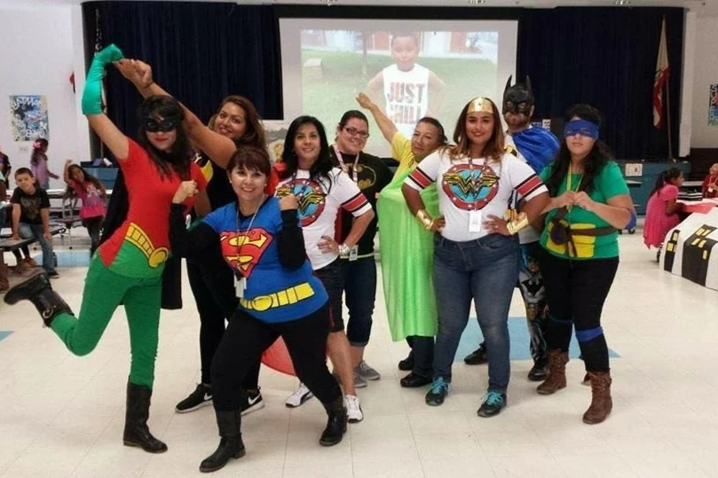 Group of nine women dressed as superhero characters, including Wonder Woman, Supergirl, Batman, and Robin, posing in a decorated indoor space with children in the background and a large screen displaying a child on stage.