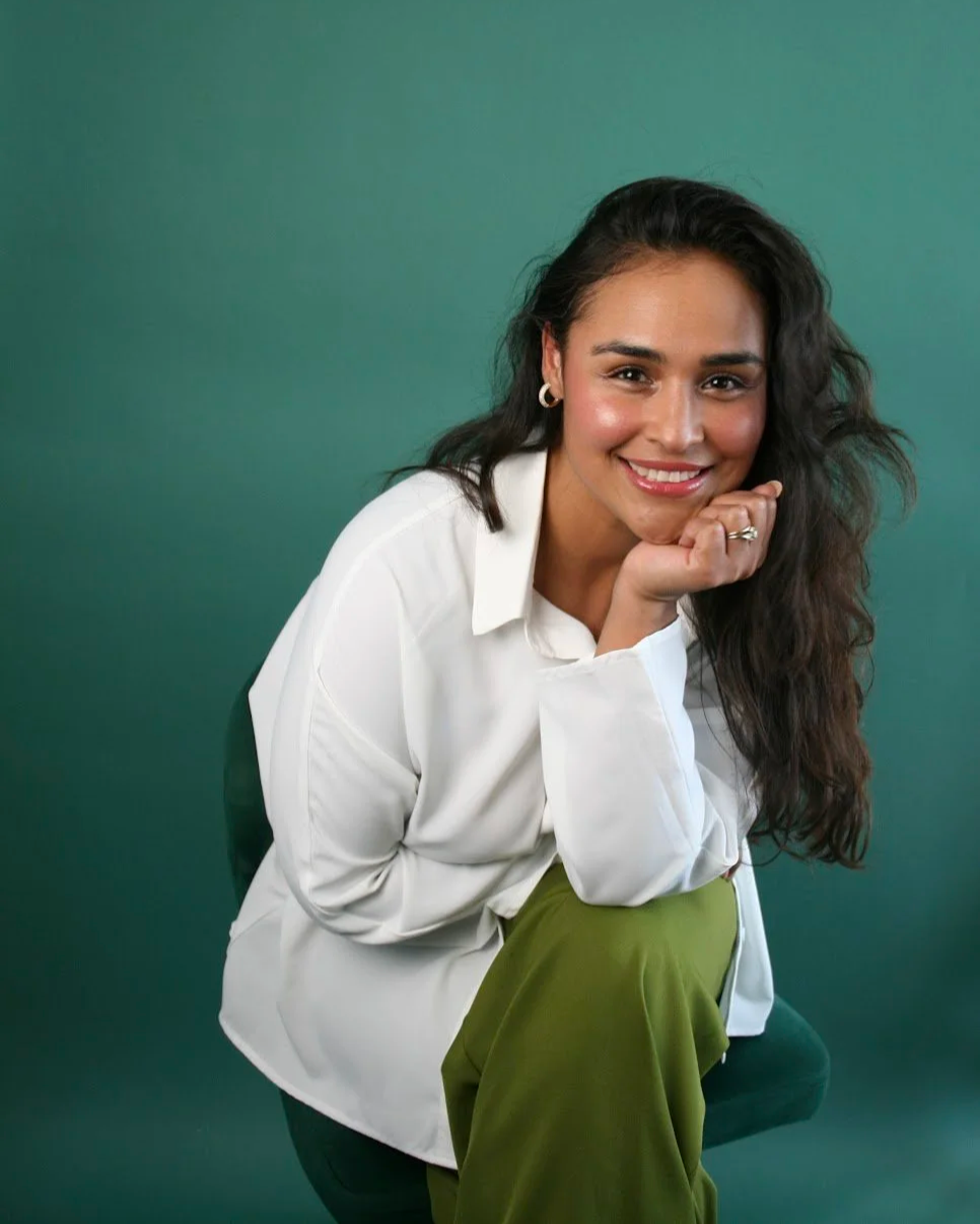 A woman with long dark wavy hair, wearing a white shirt and green pants, sitting on a stool against a green background, smiling and resting her chin on her hand.