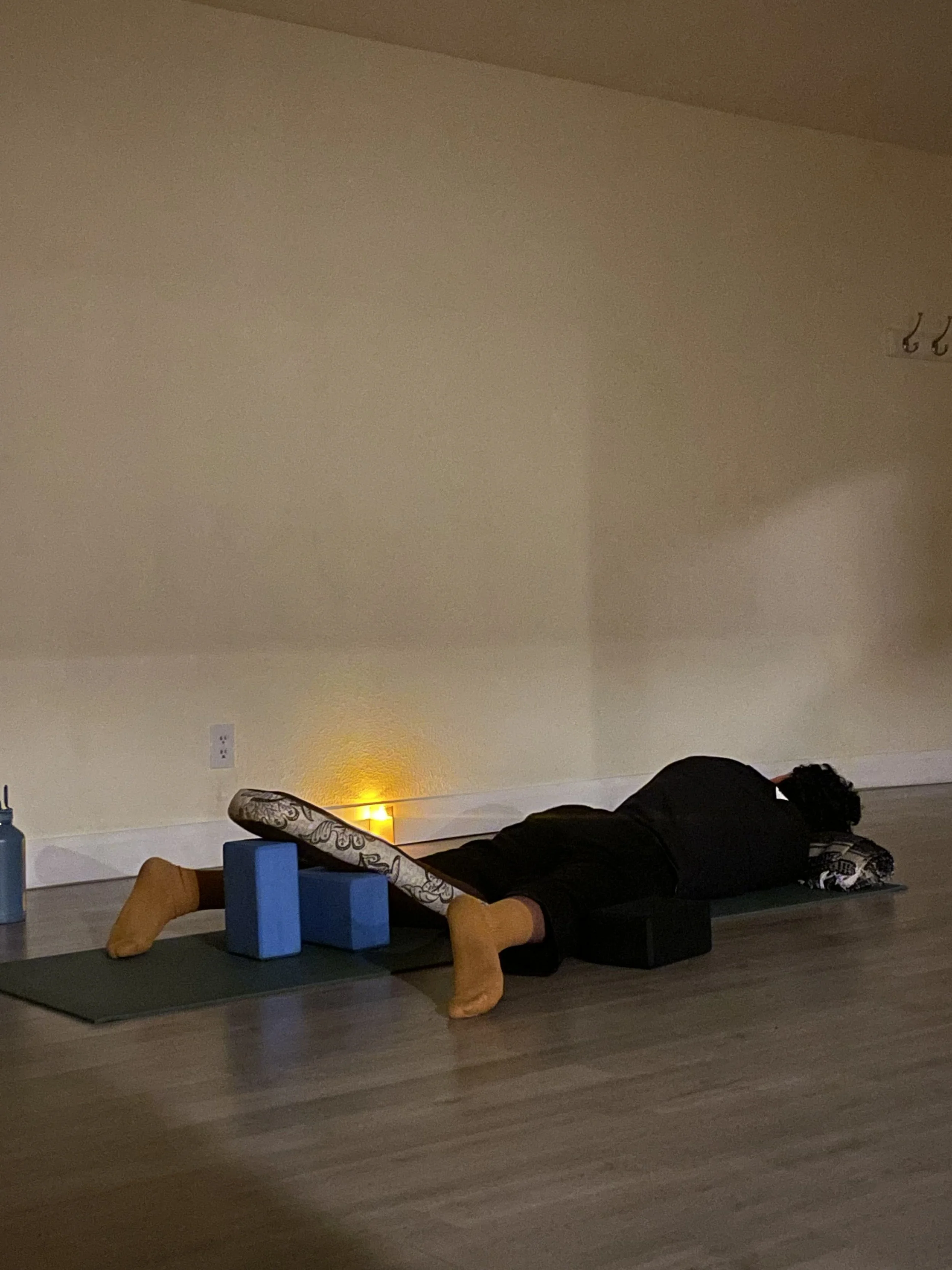 Person lying face down on a yoga mat, wearing yellow socks, with yoga blocks, a bolster, and a blanket nearby in a room with beige walls and a small lamp.