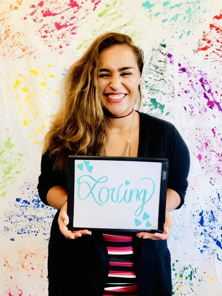 A woman with long, wavy hair smiling and holding a whiteboard that says 'Loving' with teal writing and hearts, standing against a colorful, splattered paint wall.