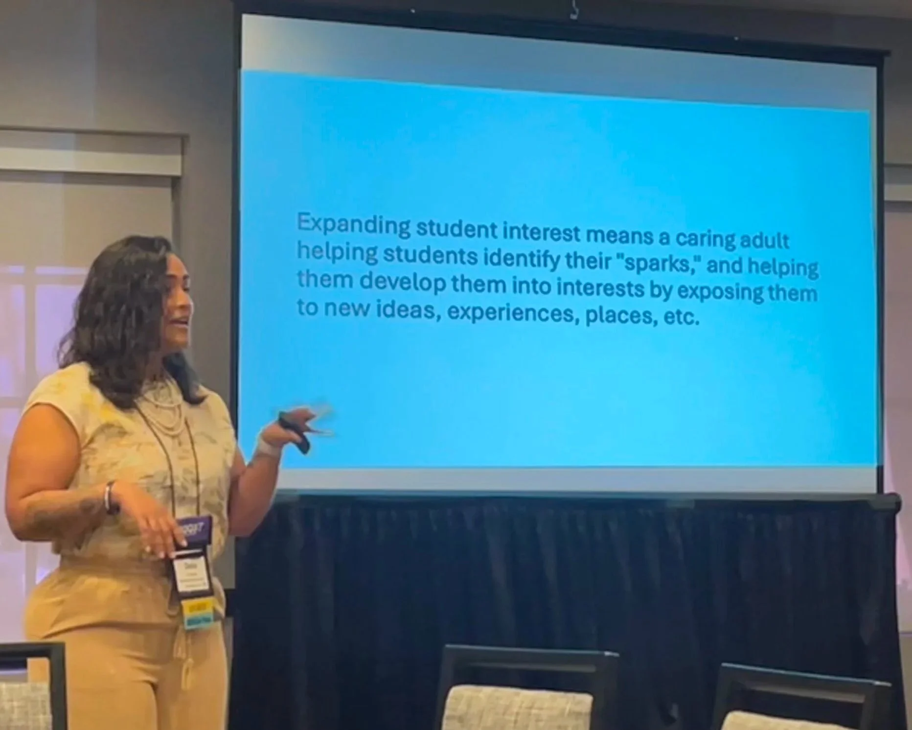 A woman in yellow attire presenting in front of a large screen displaying a slide about expanding student interest, with text explaining that caring adults help students identify their sparks and develop interests through exposure.