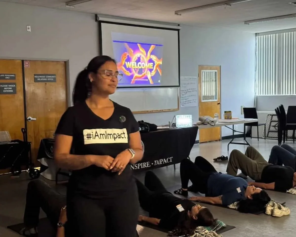 A woman standing in front of a classroom of people lying on mats on the floor during a workshop or seminar, with a welcome message on a screen behind her.