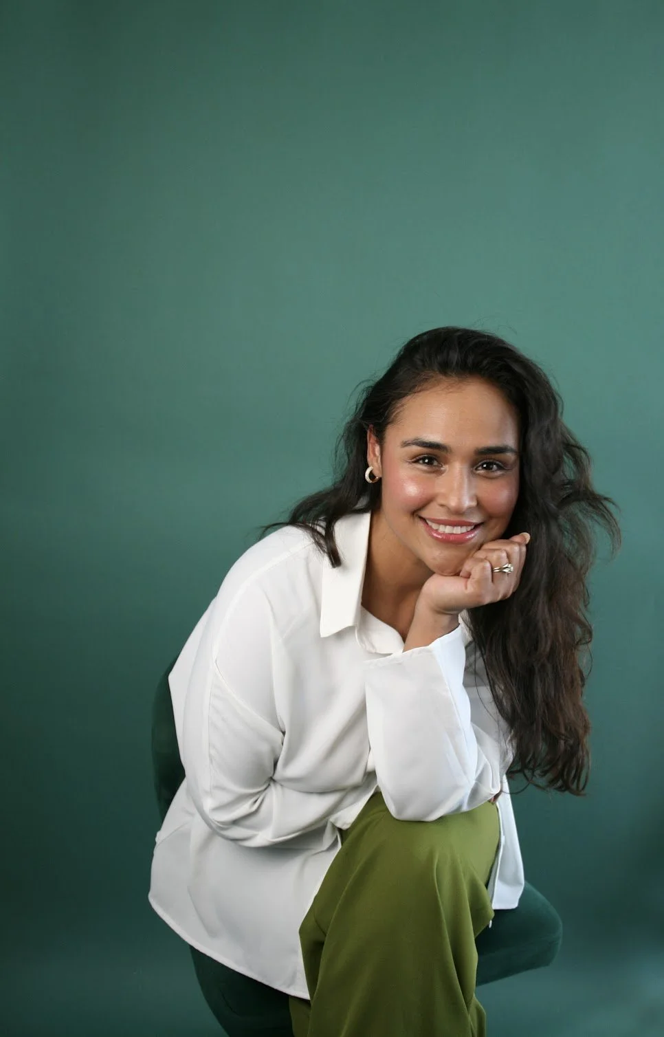 A woman with long wavy hair smiling, wearing a white blazer and green pants, seated against a green background.