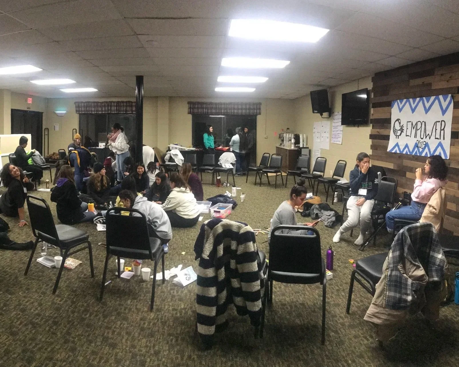 Group of people gathered in a large room with chairs, tables, and a banner reading 'EMPOWER 2019' on the wall. Some people are sitting on the floor, others are standing and talking, and a few are sitting and interacting.