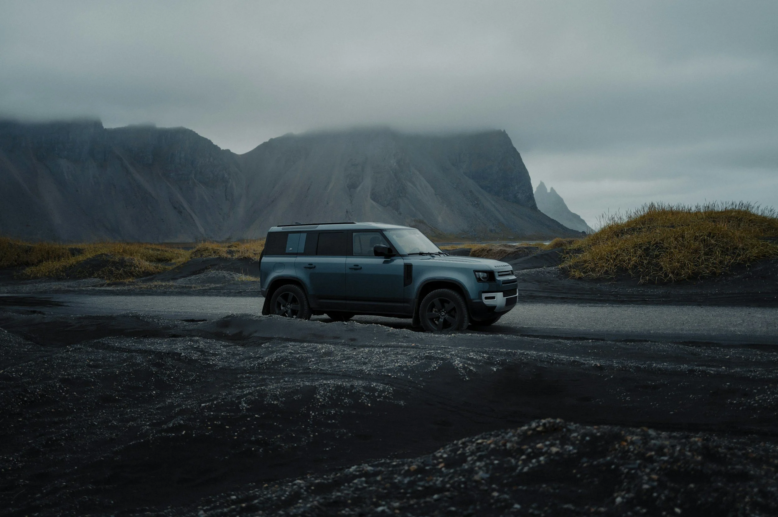 A blue off-road vehicle on a rugged, dark terrain with mountains and cloudy sky in the background