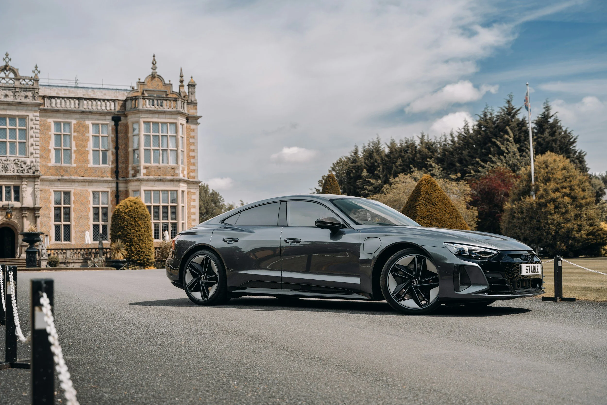 A sleek black modern sports car parked in front of a stately historic building with a garden and trees
