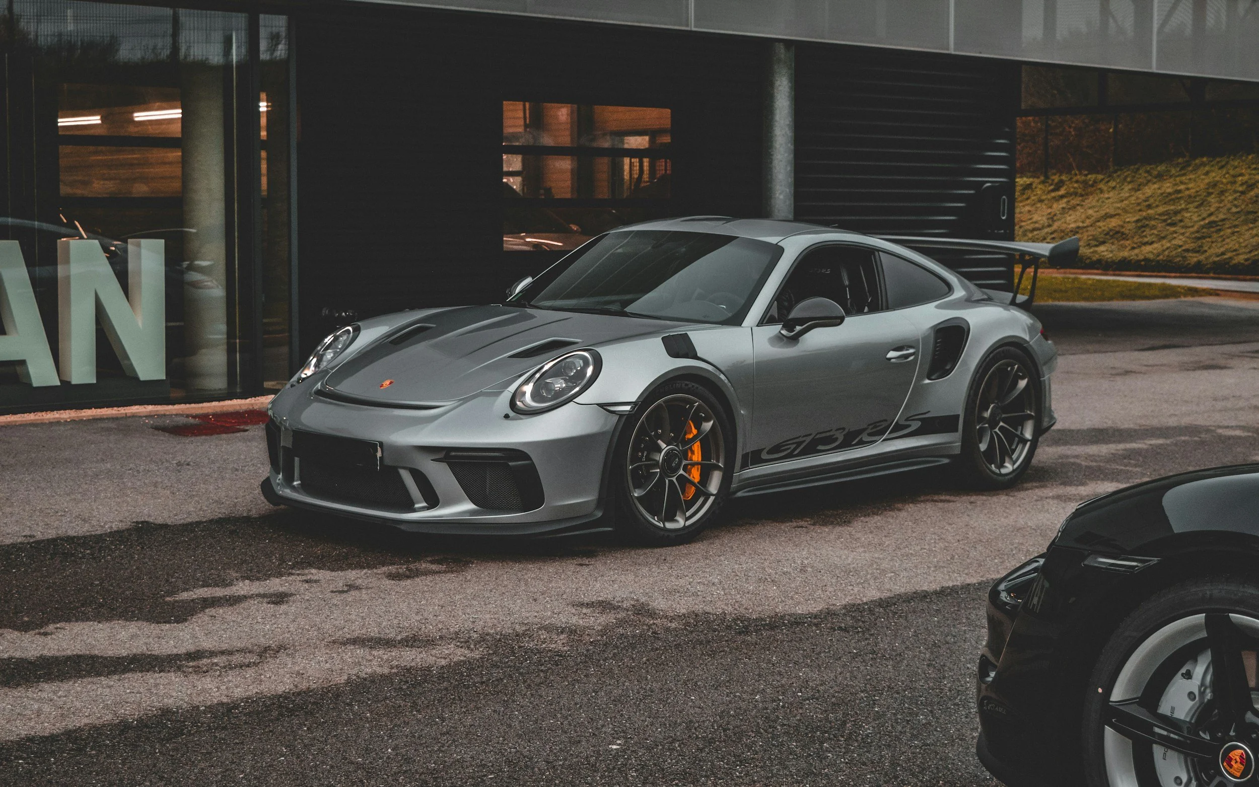 A silver Porsche 911 GT3 RS sports car parked outside a modern building with large glass windows.
