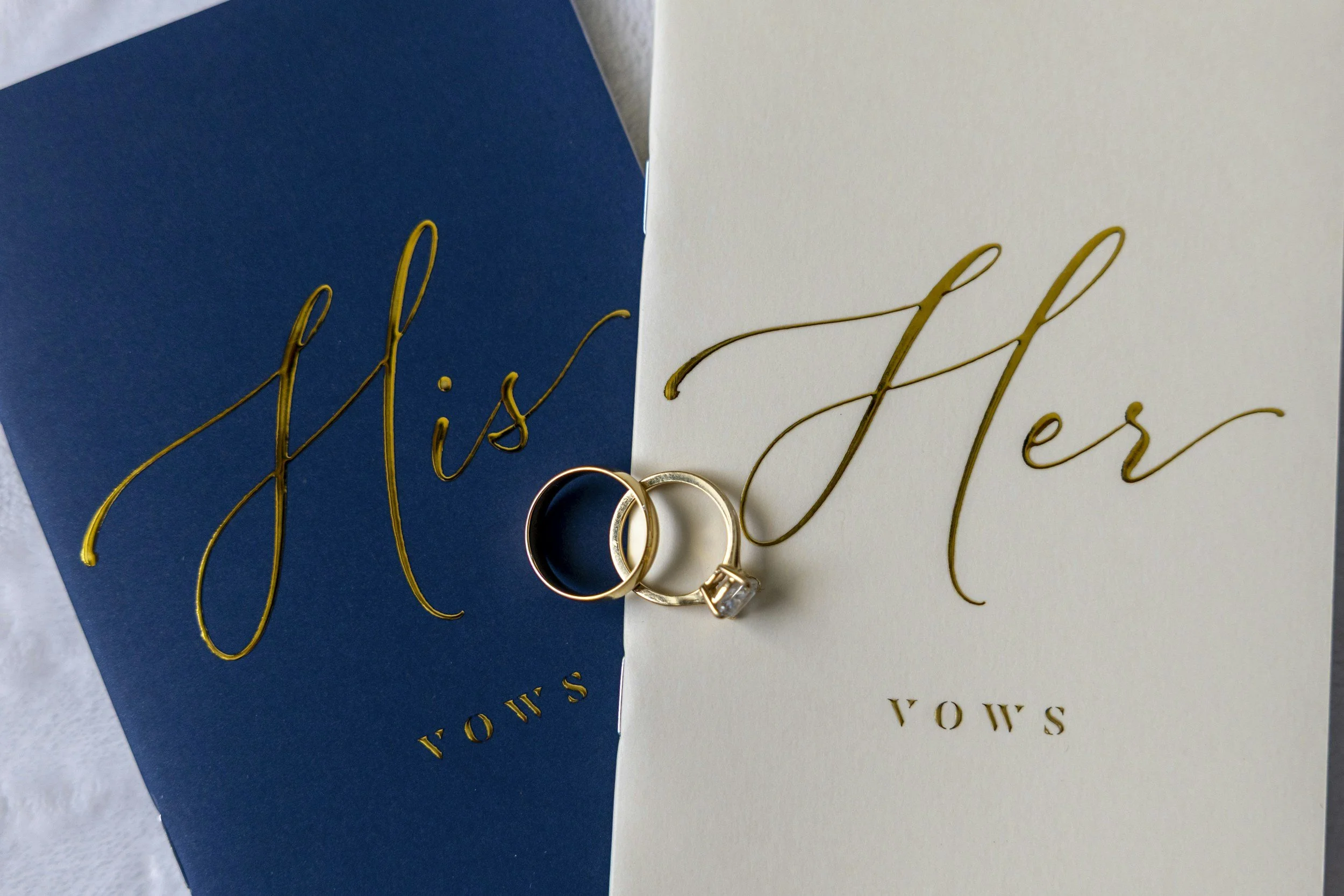 Close-up view of two wedding rings placed on top of wedding vows cards; one card is dark blue and the other is white, both with gold lettering.