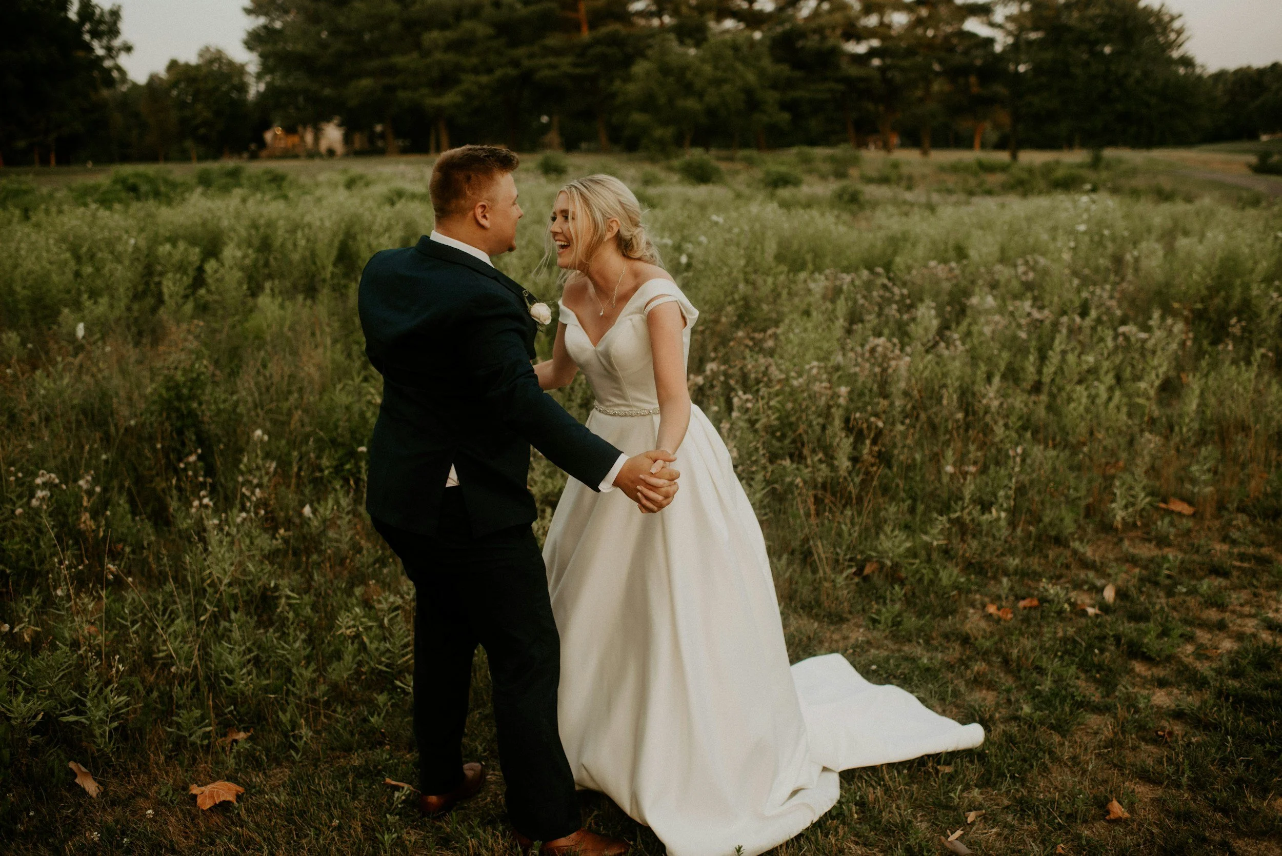 A bride and groom share a dance outdoors in a field with green plants, trees, and a distant house in the background during sunset, with the bride wearing a white wedding dress and the groom in a dark suit.