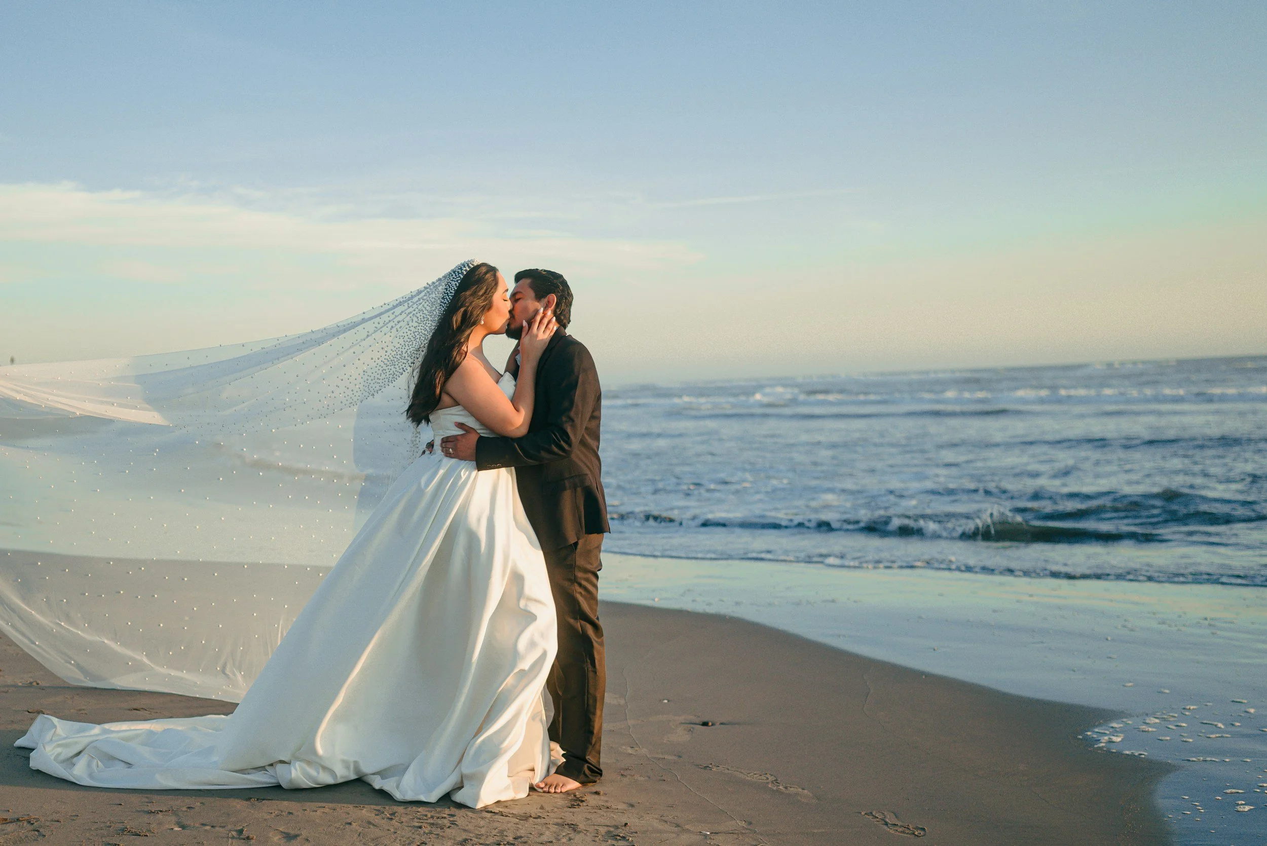 A bride and groom share a kiss on the beach, with the bride in a white wedding gown and veil, and the groom in a black suit. The ocean waves are in the background, and the bride's veil flows in the wind.