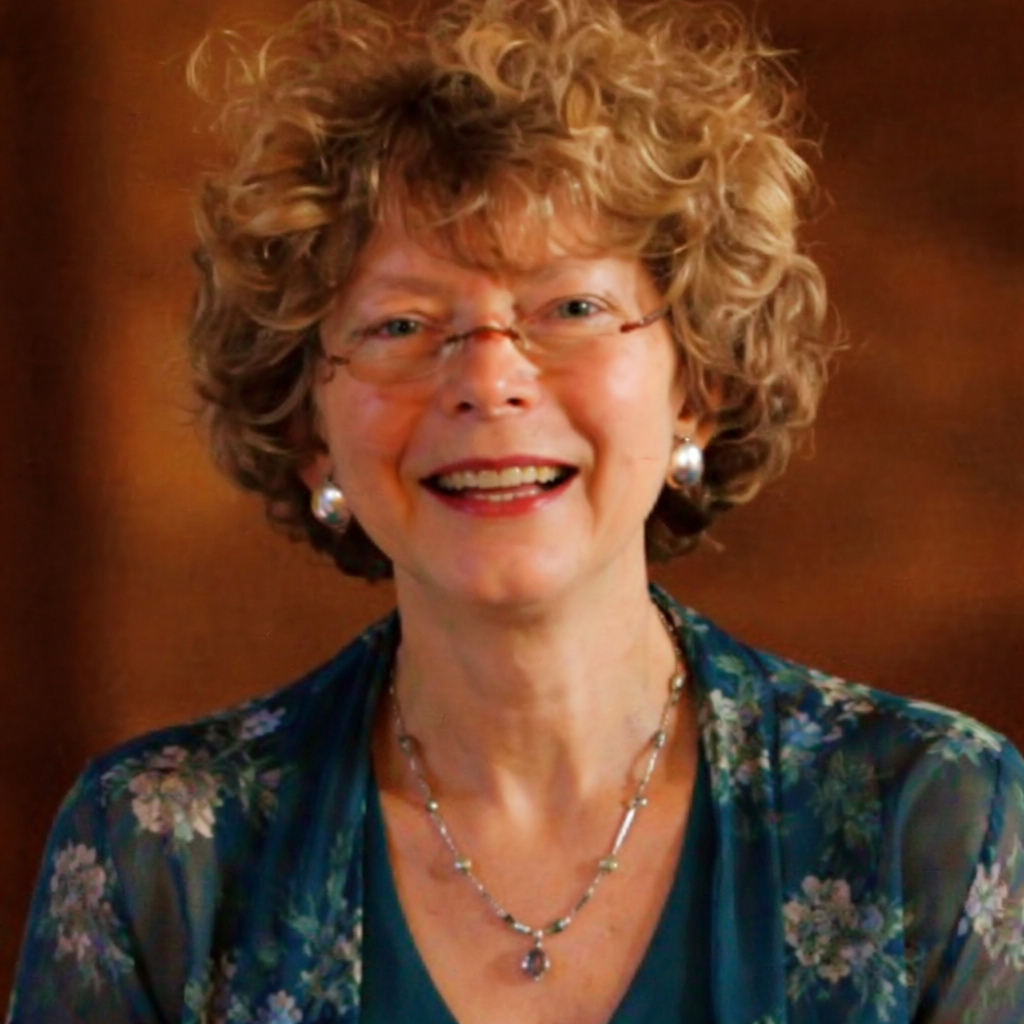 A smiling woman with curly blonde hair and glasses, wearing a green floral top and jewelry, posing against a warm, brown background.