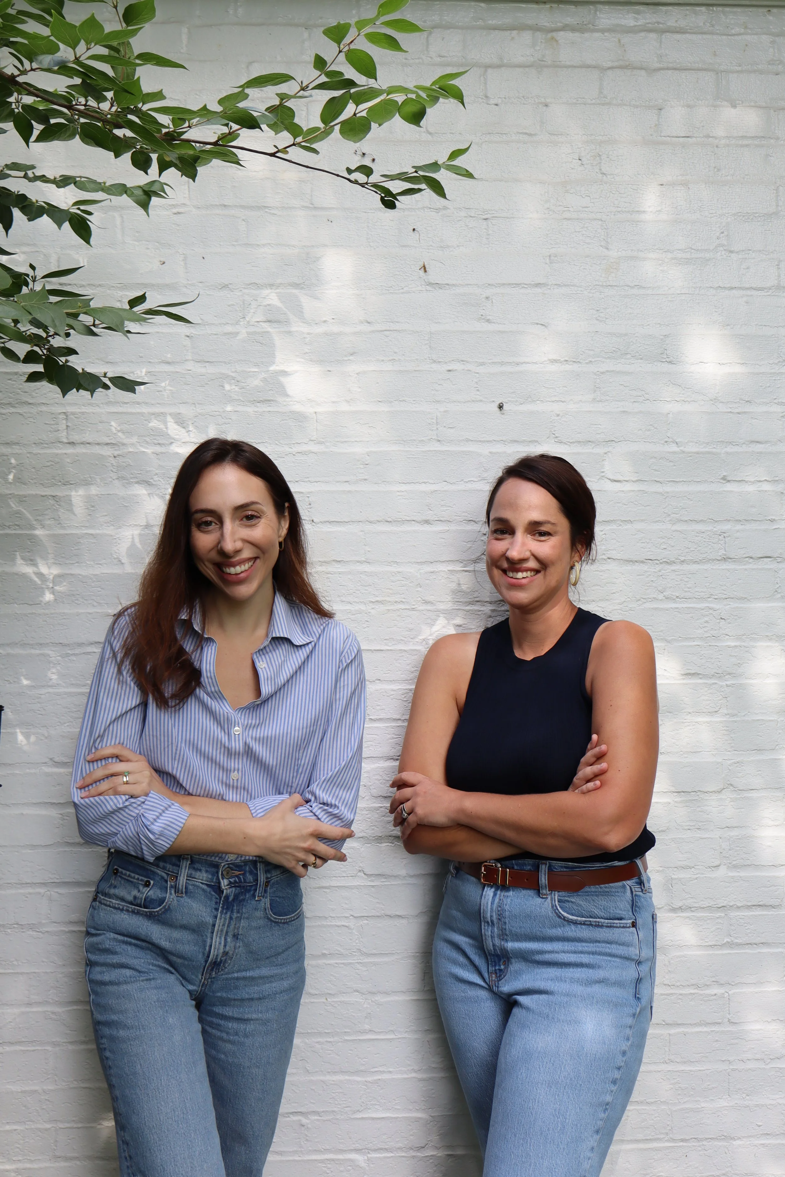 Two women standing against a white brick wall, smiling, with arms crossed, one wearing a blue striped shirt and jeans, the other in a black tank top and jeans.