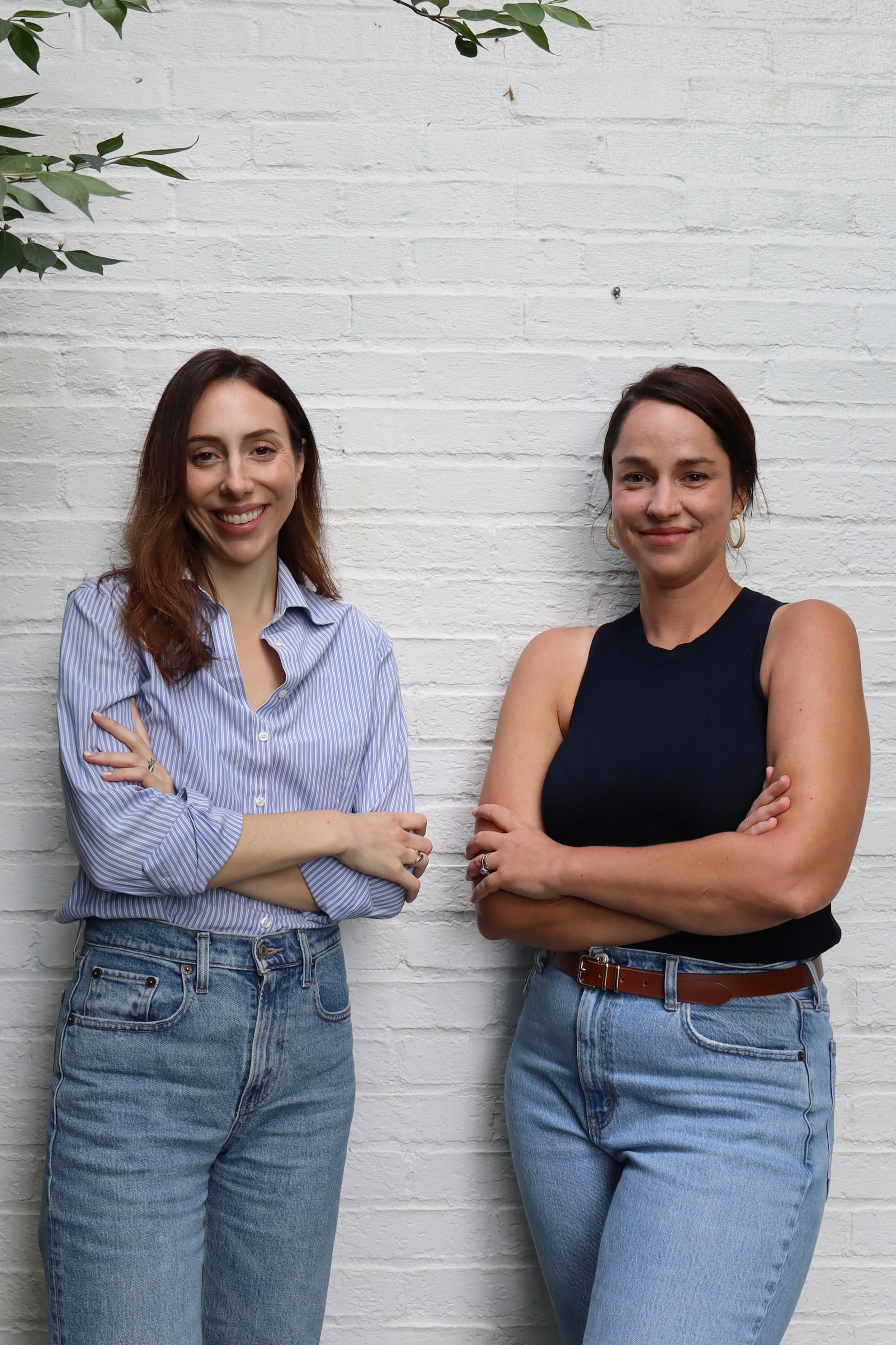 Two women standing with arms crossed in front of a white brick wall, smiling. The woman on the left has long, wavy brown hair, is wearing a blue and white striped button-up shirt and high-waisted blue jeans. The woman on the right has short dark hair, is wearing hoop earrings and a sleeveless black top with blue jeans.