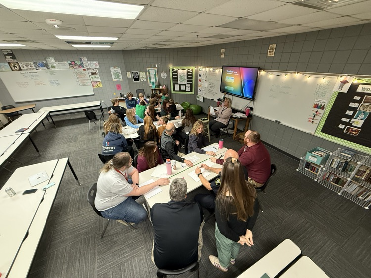A classroom with students and adults sitting around tables, attending a meeting or discussion in a school setting.